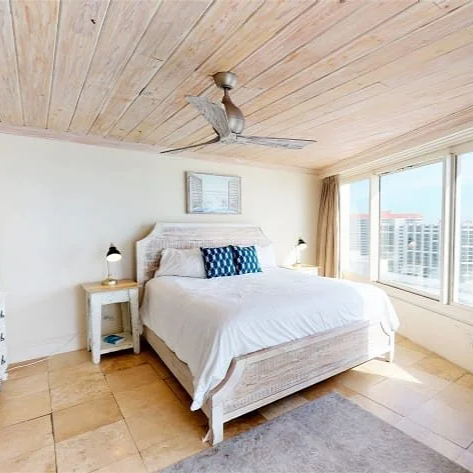 Bright bedroom with a white bed, nightstands, lamps, and a ceiling fan, overlooking city buildings through large windows.