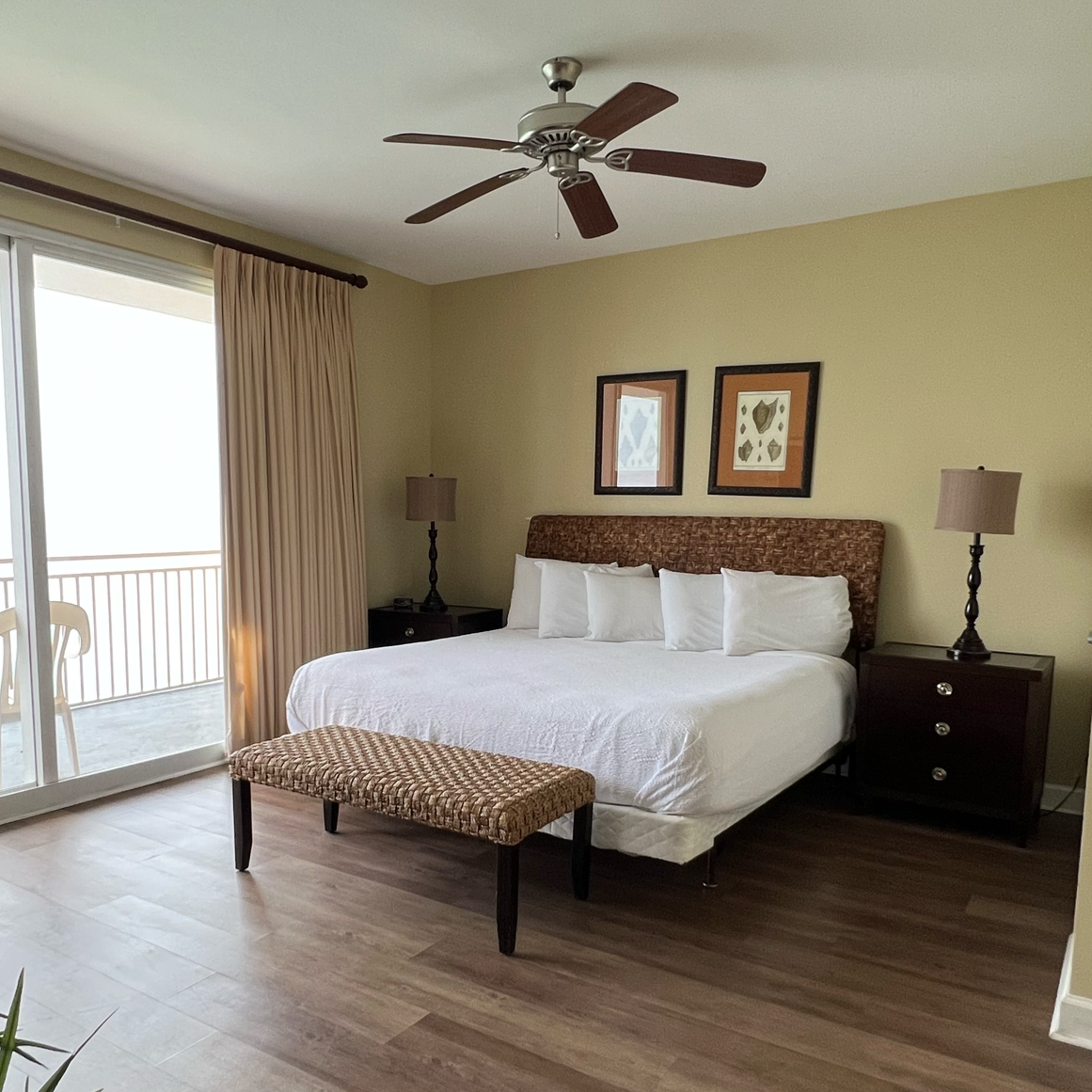 A hotel room with a large bed, white linens, multiple pillows, and a woven headboard. Two dark wood nightstands with lamps, framed artwork above the bed, a ceiling fan, and a sliding glass door with beige curtains leading to a balcony with a chair.