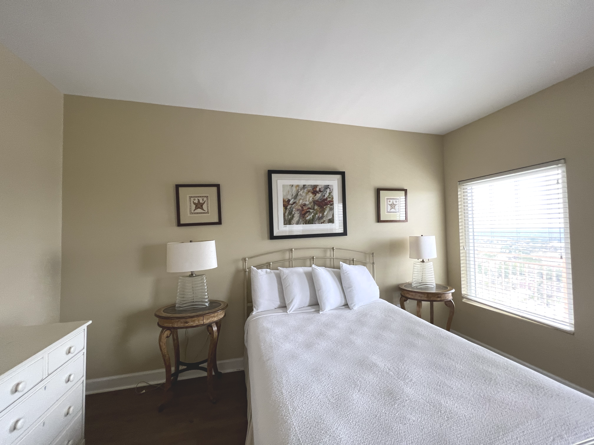 A bedroom with a bed, white bedding, four pillows, two bedside tables with lamps, artwork on the beige wall, and a window with blinds letting in natural light.