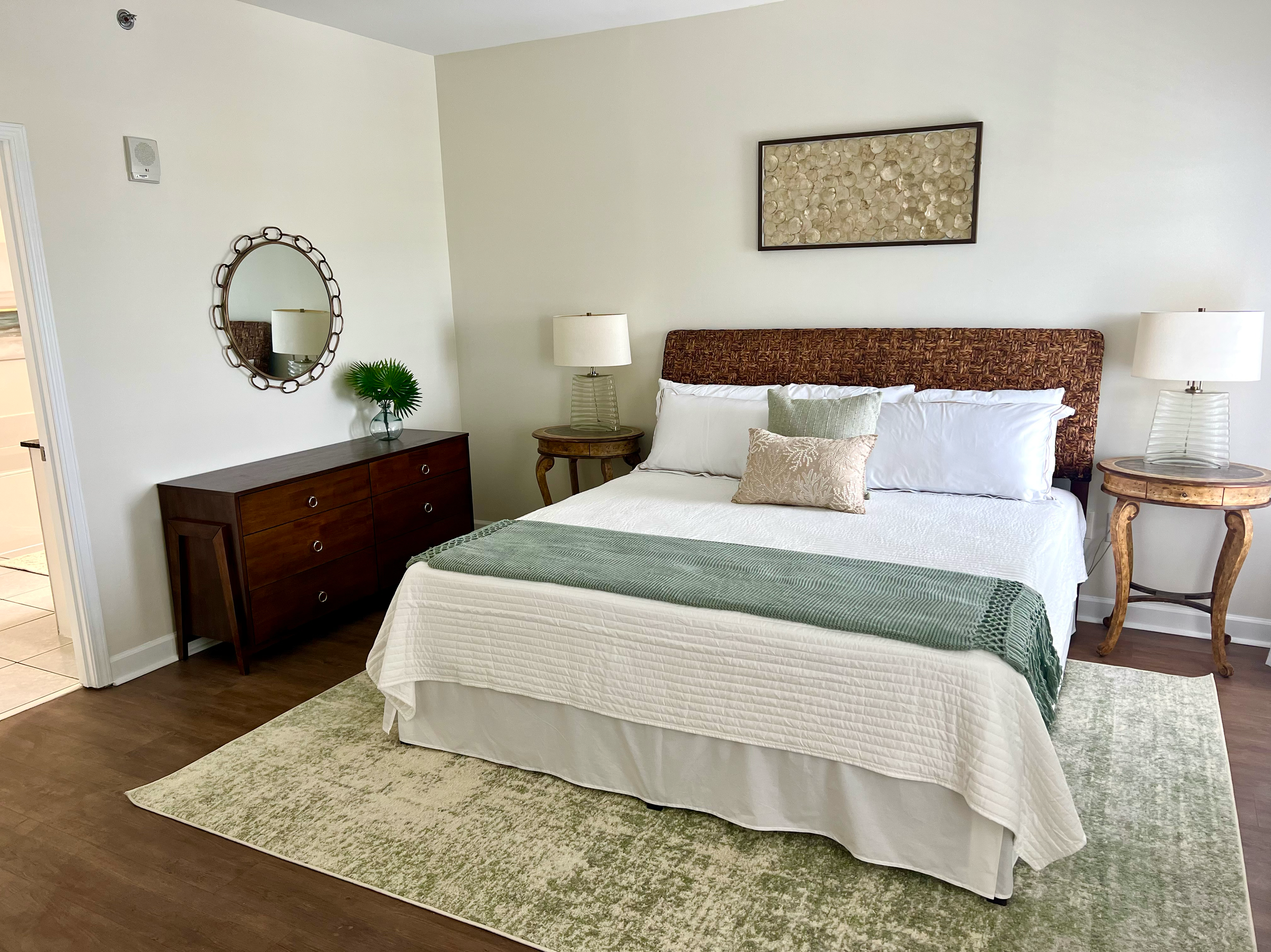 A neatly made bed with white bedding, green throw, and decorative pillows, positioned between two wooden nightstands with lamps. A patterned headboard is behind the bed, and a framed artwork hangs above. A dark wood dresser with a mirror and a small