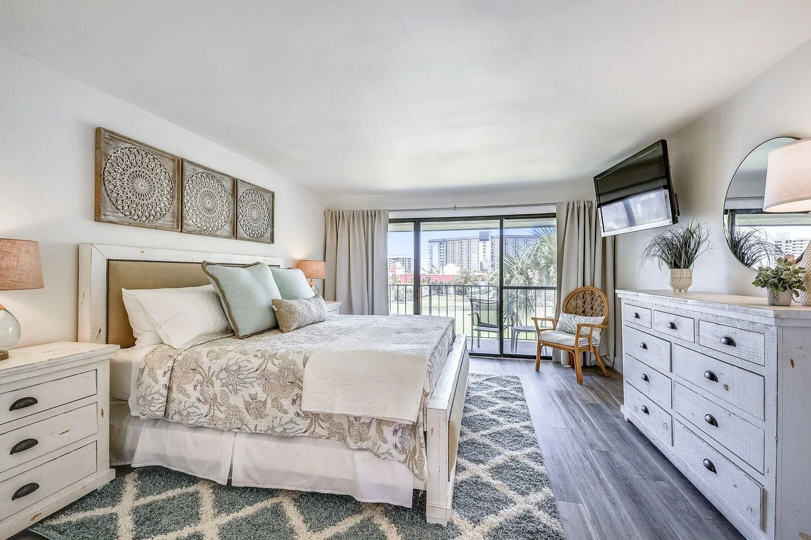 Bright bedroom with a large bed, white wooden furniture, and a sliding glass door leading to a balcony with outdoor seating, city view, and tropical plants.