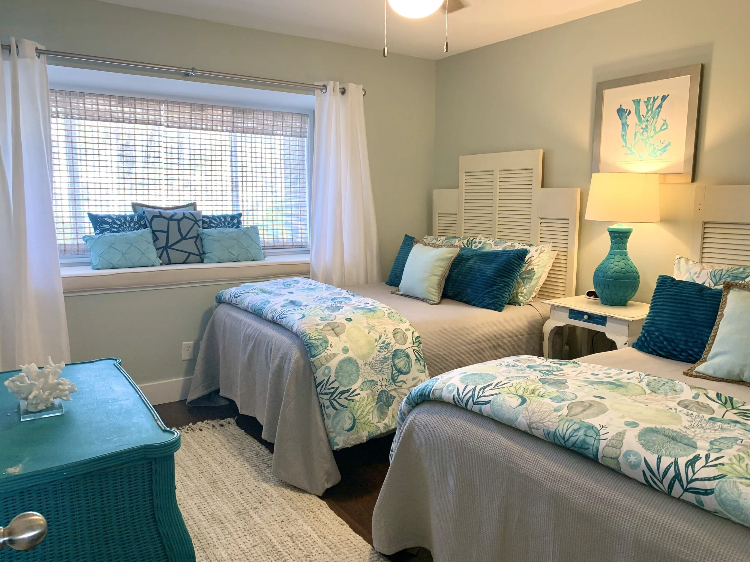 A bedroom with beach-themed decor featuring two beds with seashell and coral-patterned bedding in shades of blue, green, and white. There is a light-colored nightstand with a turquoise lamp, a window with white curtains and a window seat decorated wi