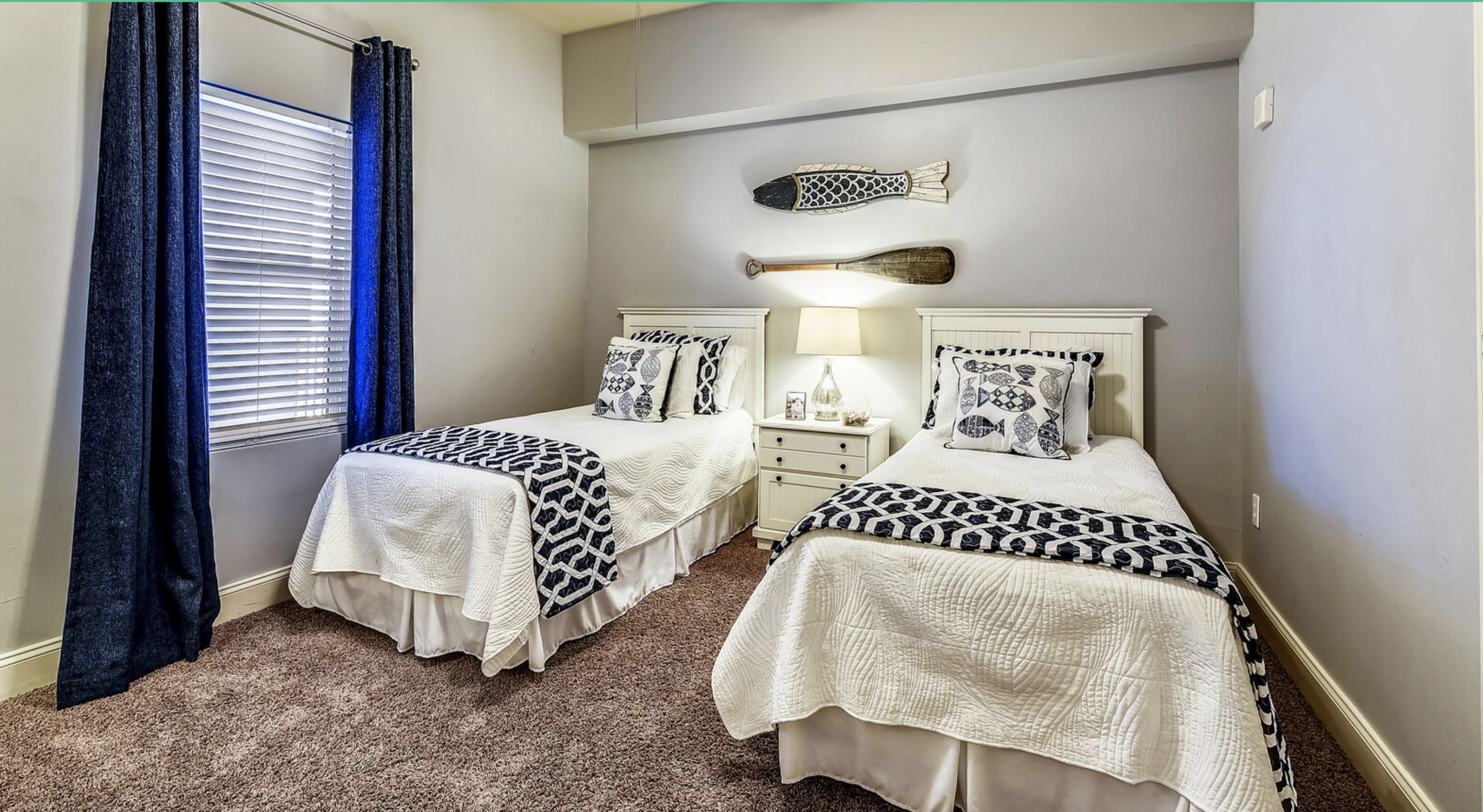 A bedroom with two twin beds, nautical-themed bedding, a nightstand with a lamp, and wall decorations including a fish and oar, with a window covered by blue curtains.