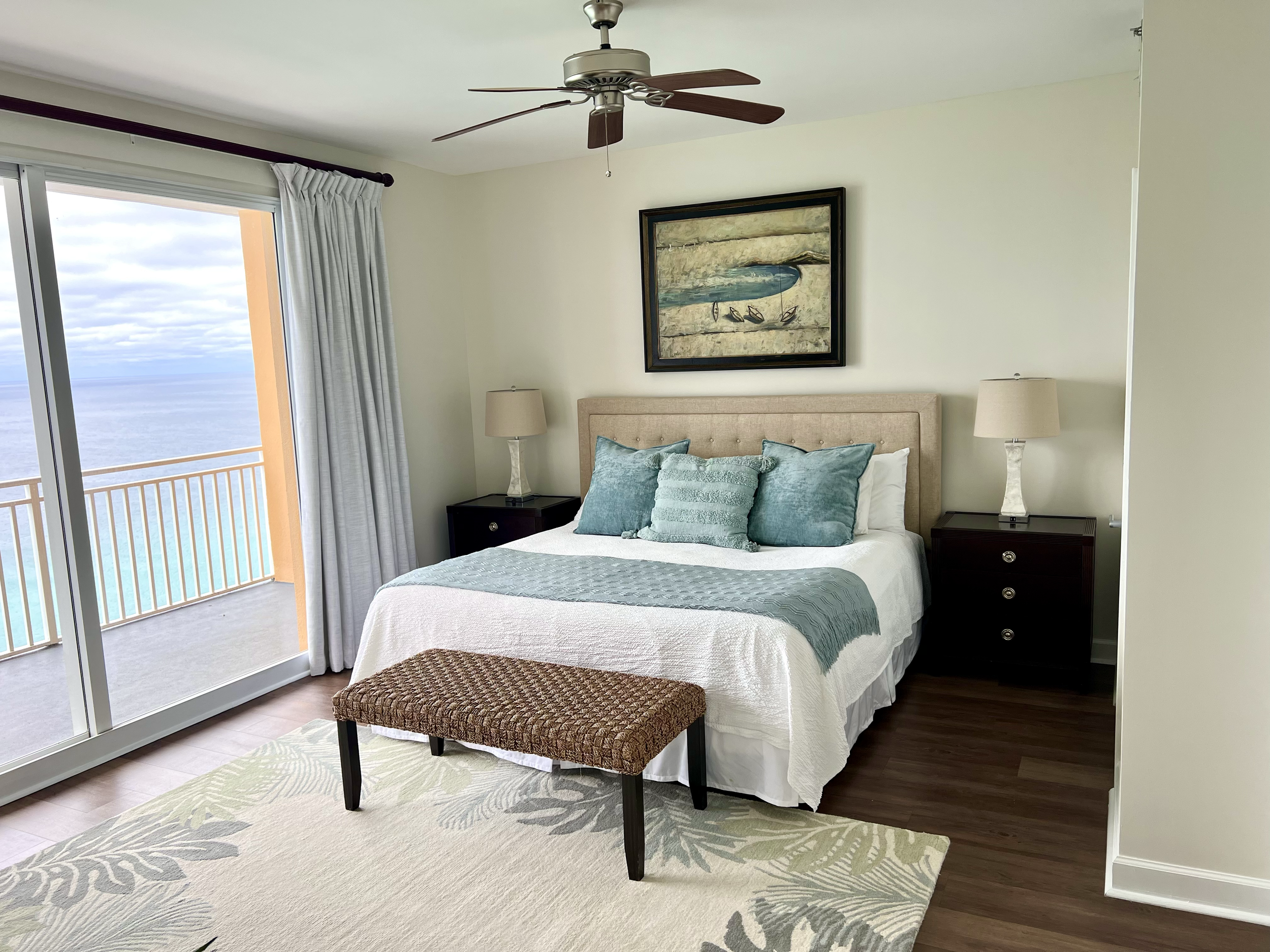 Beach view from a bedroom with a sliding glass door, a bed with teal pillows and a bench at the foot, and two nightstands with lamps.