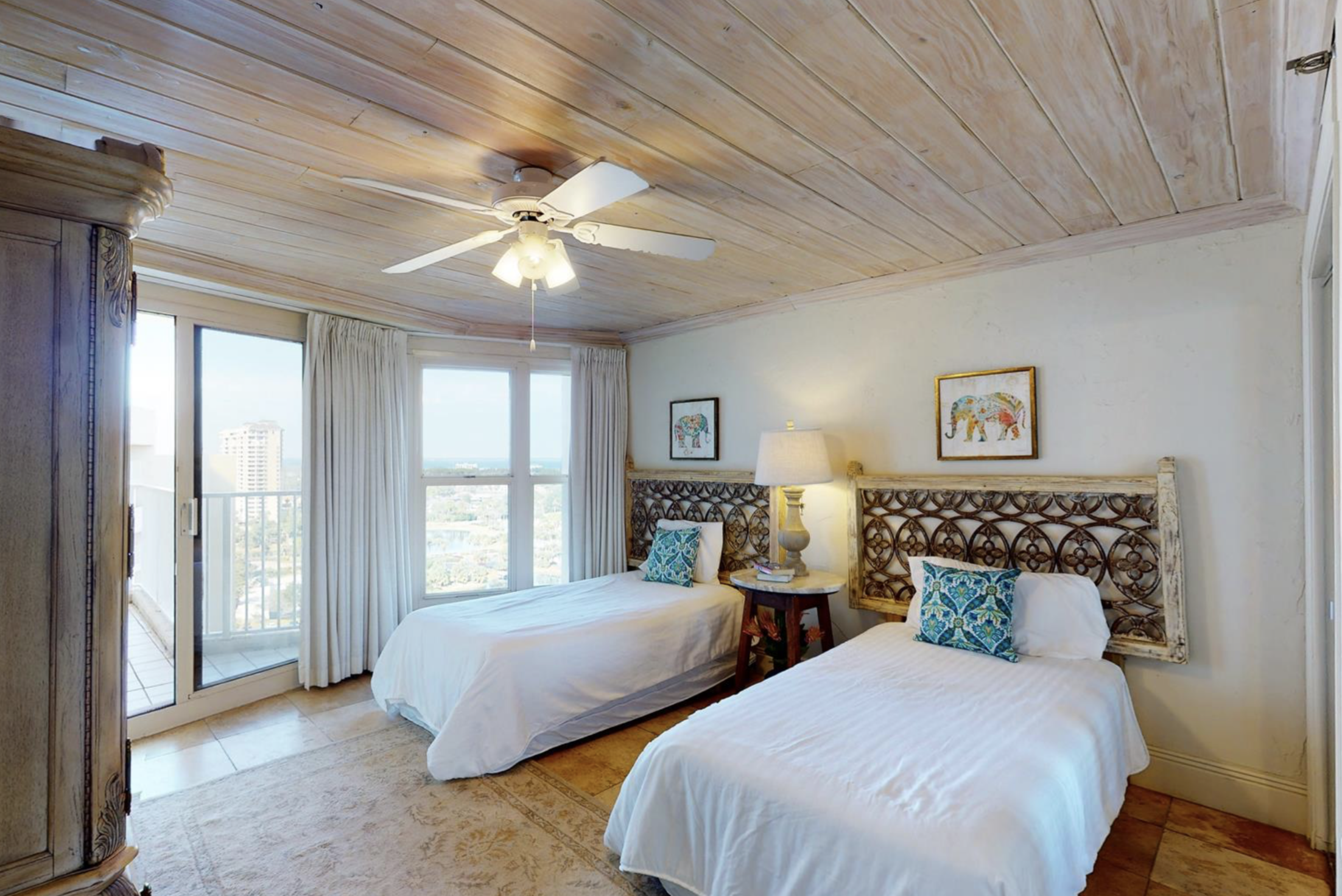 Bedroom with two twin beds, wooden headboards, nightstand with lamp, framed elephant artwork, large window with white curtains, ceiling fan, and wooden ceiling.