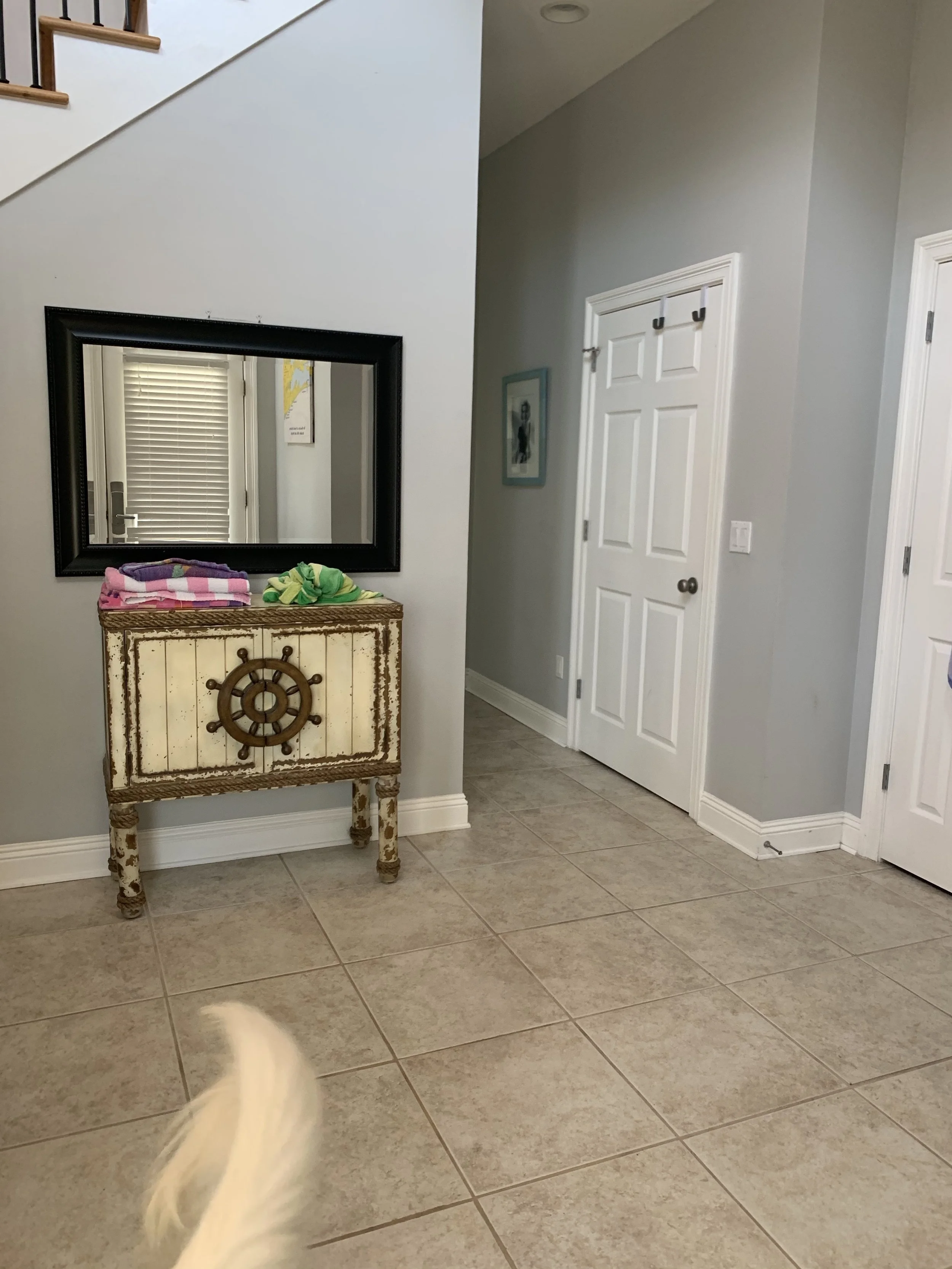 Entryway with tiled floor, white door, wall hooks, a framed picture, a mirror, and a decorative cabinet with sea-themed design, with towels on top.