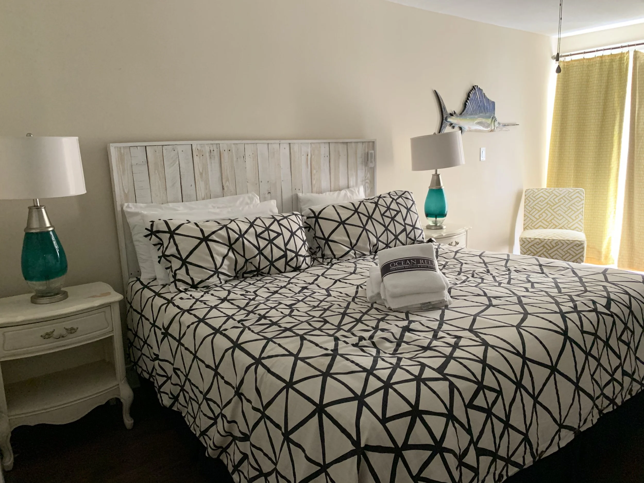 A bedroom with a white wooden headboard, two nightstands with turquoise lamps, a patterned bedspread, a chair, and a fish wall sculpture by the window with yellow curtains.