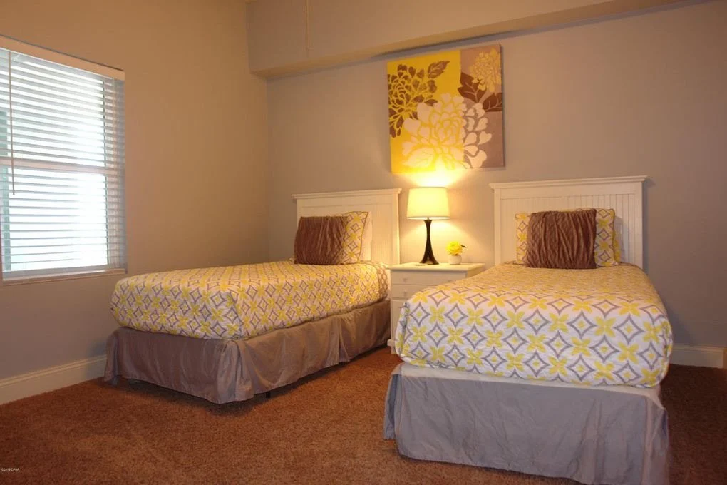 A bedroom with two twin beds with floral bedding and brown accent pillows, a white nightstand with a lamp and small flower, a window with blinds, beige wall art above the beds, and brown carpet.