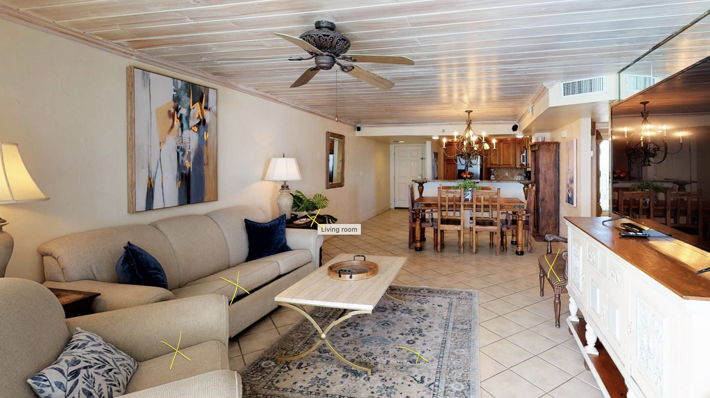 Spacious living room and open kitchen area in a home with beige walls, wooden ceiling, ceiling fan, art on the wall, beige sofa with navy cushions, a coffee table, a patterned area rug, a large flat-screen TV on a white entertainment console, and a d
