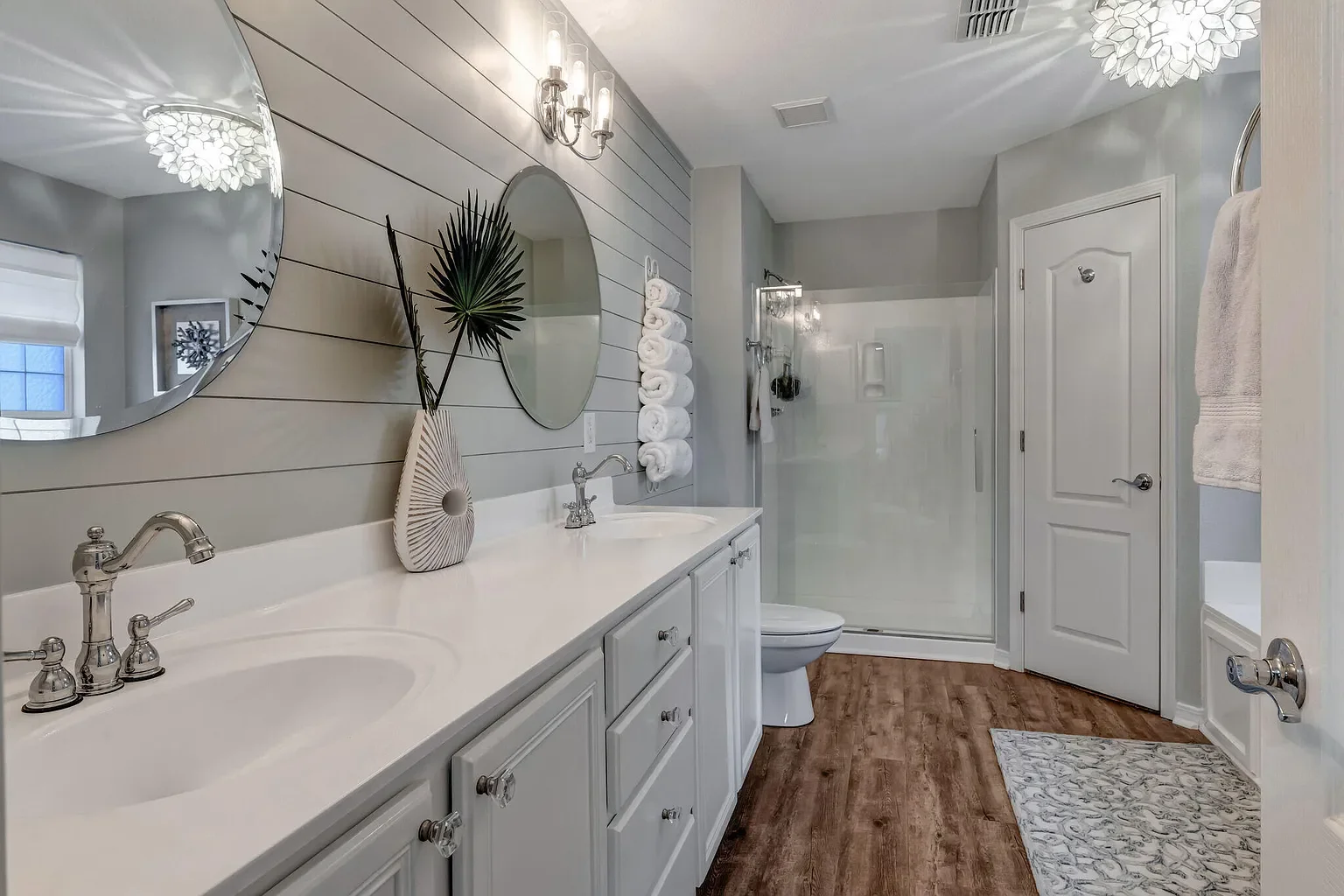 A modern bathroom with a double vanity, two oval mirrors, a decorative vase with a palm leaf, and rolled towels on a rack. It features a glass shower enclosure, wood-look flooring, and light gray shiplap walls.