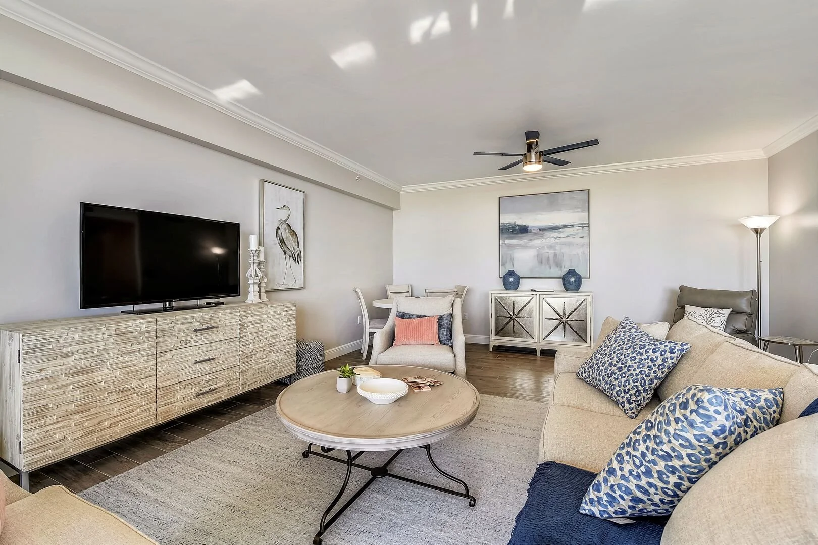 Living room with a beige sofa, patterned pillows, a round coffee table, a flat-screen TV on a textured white TV stand, wall art, a white armchair with decorative pillows, a white sideboard with blue vases, a floor lamp, and a ceiling fan.