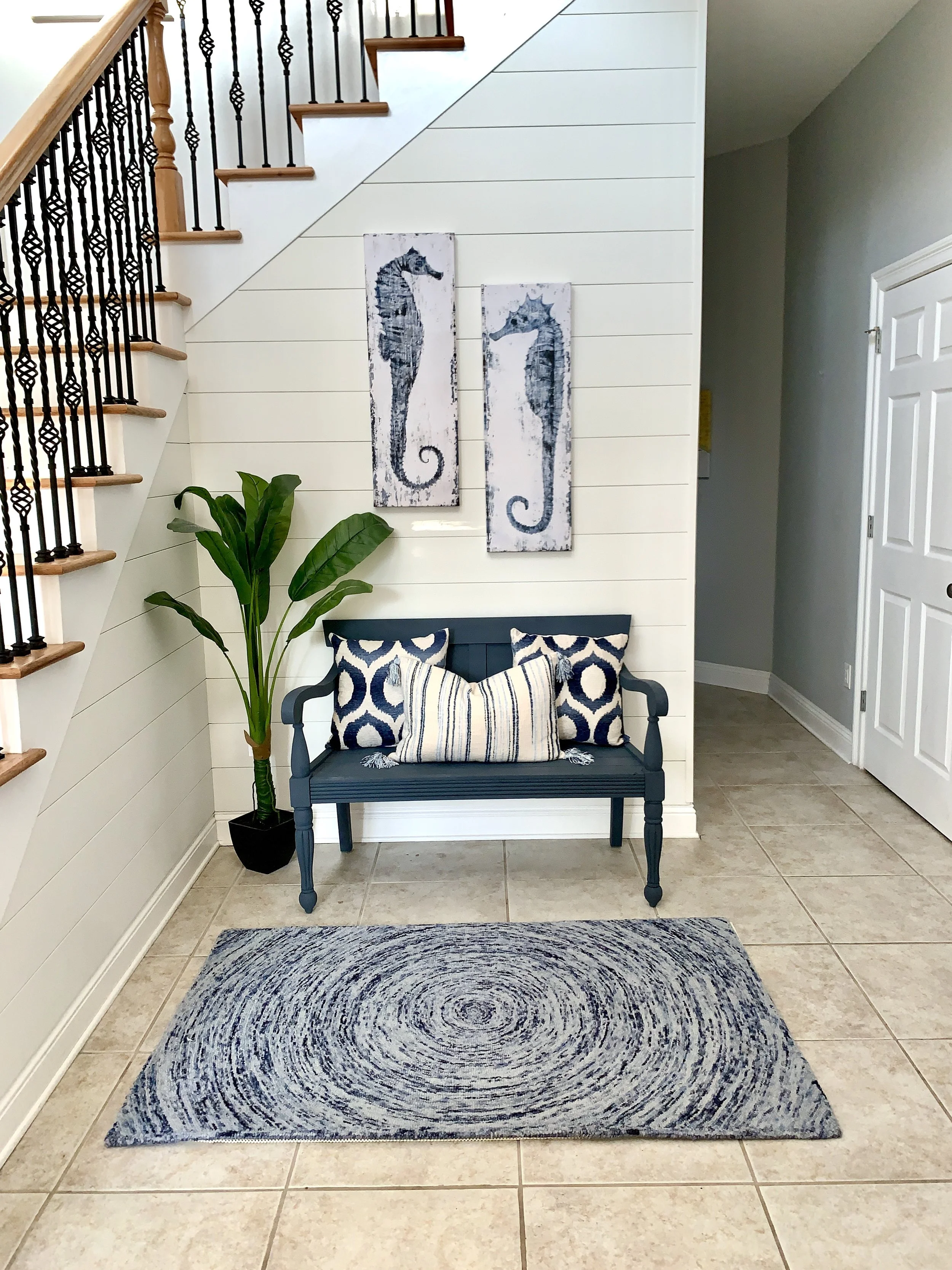 Entryway with a navy blue bench, patterned pillows, potted plant, two seahorse wall art pieces, beige tiled floor, white shiplap wall, staircase with metal and wood railing, and a door to the right.