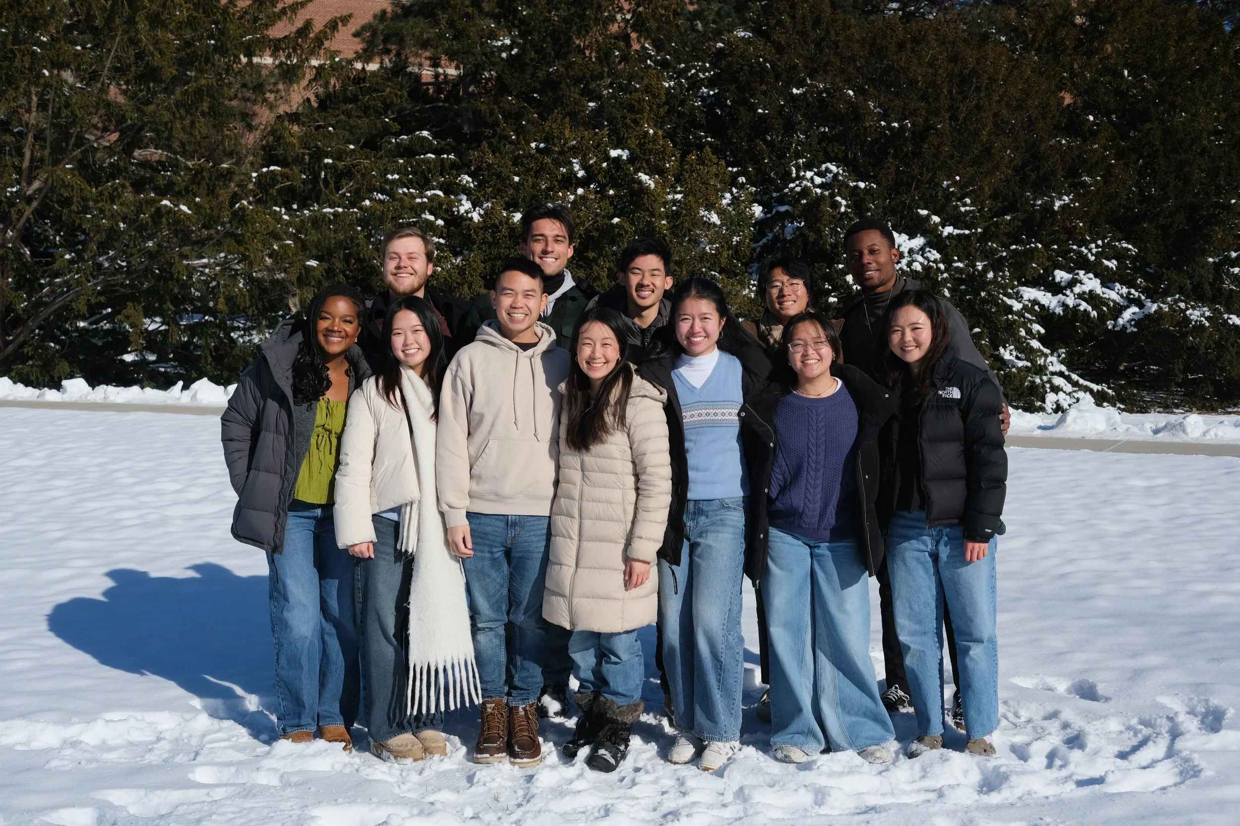 A diverse group of fourteen young adults, dressed in winter clothing, standing together outside on snow with trees in the background, smiling at the camera.