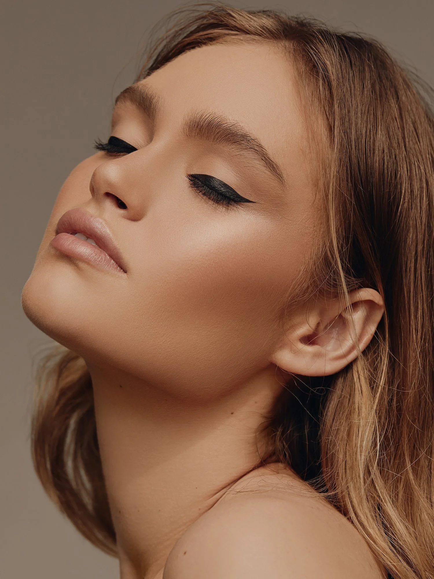 Close-up of a woman's face with makeup, featuring winged eyeliner, nude lipstick, and natural skin tone, with her eyes closed and head slightly tilted.
