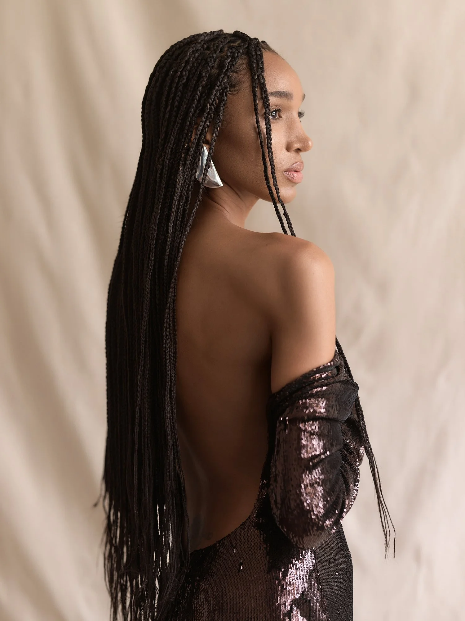 A woman with long braided hair, wearing a shiny black dress with sequins, and large silver earrings, is turned sideways with a soft beige background.