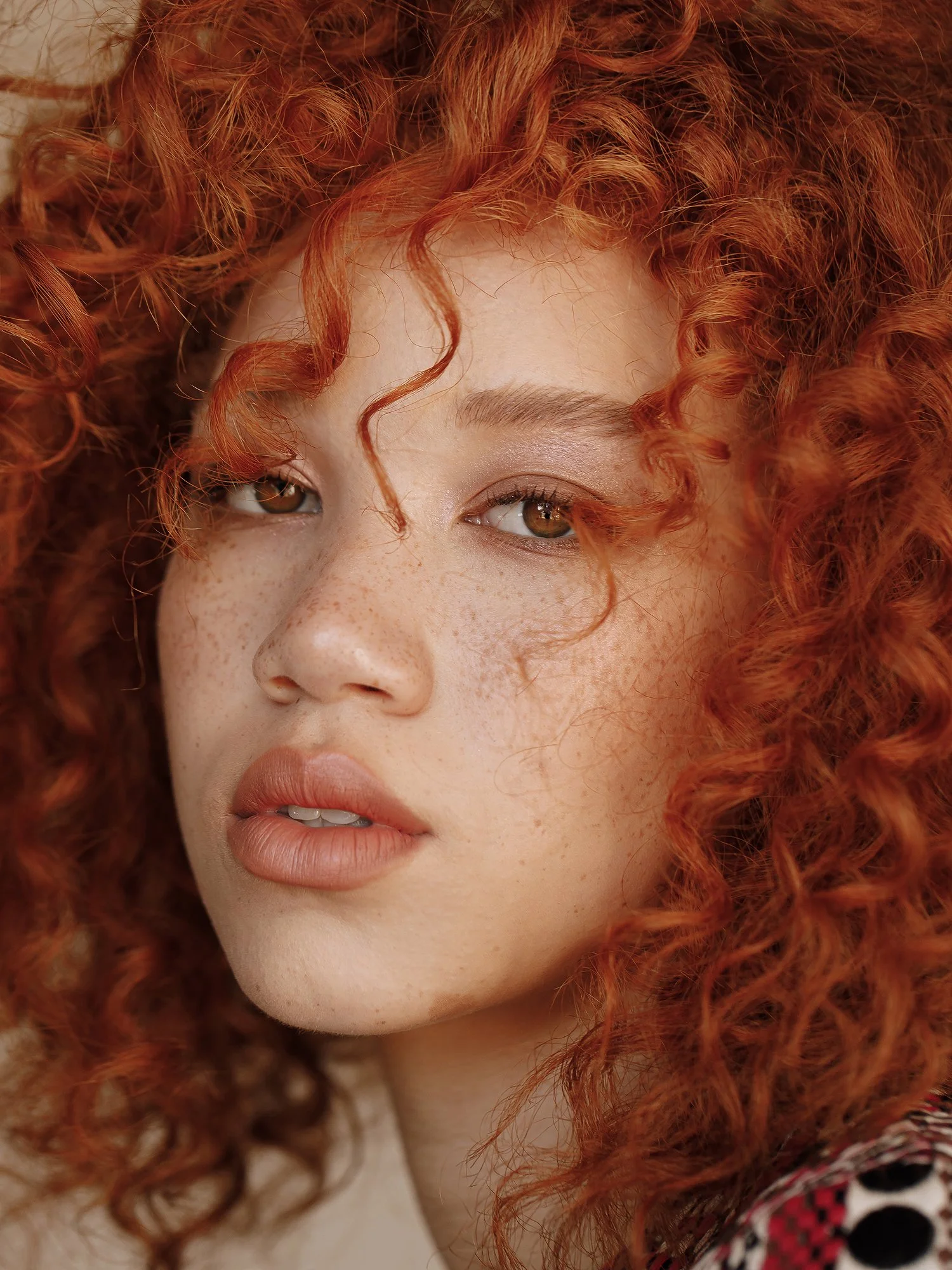 Close-up of a young woman with red curly hair, freckles, and light makeup, looking slightly to the side.