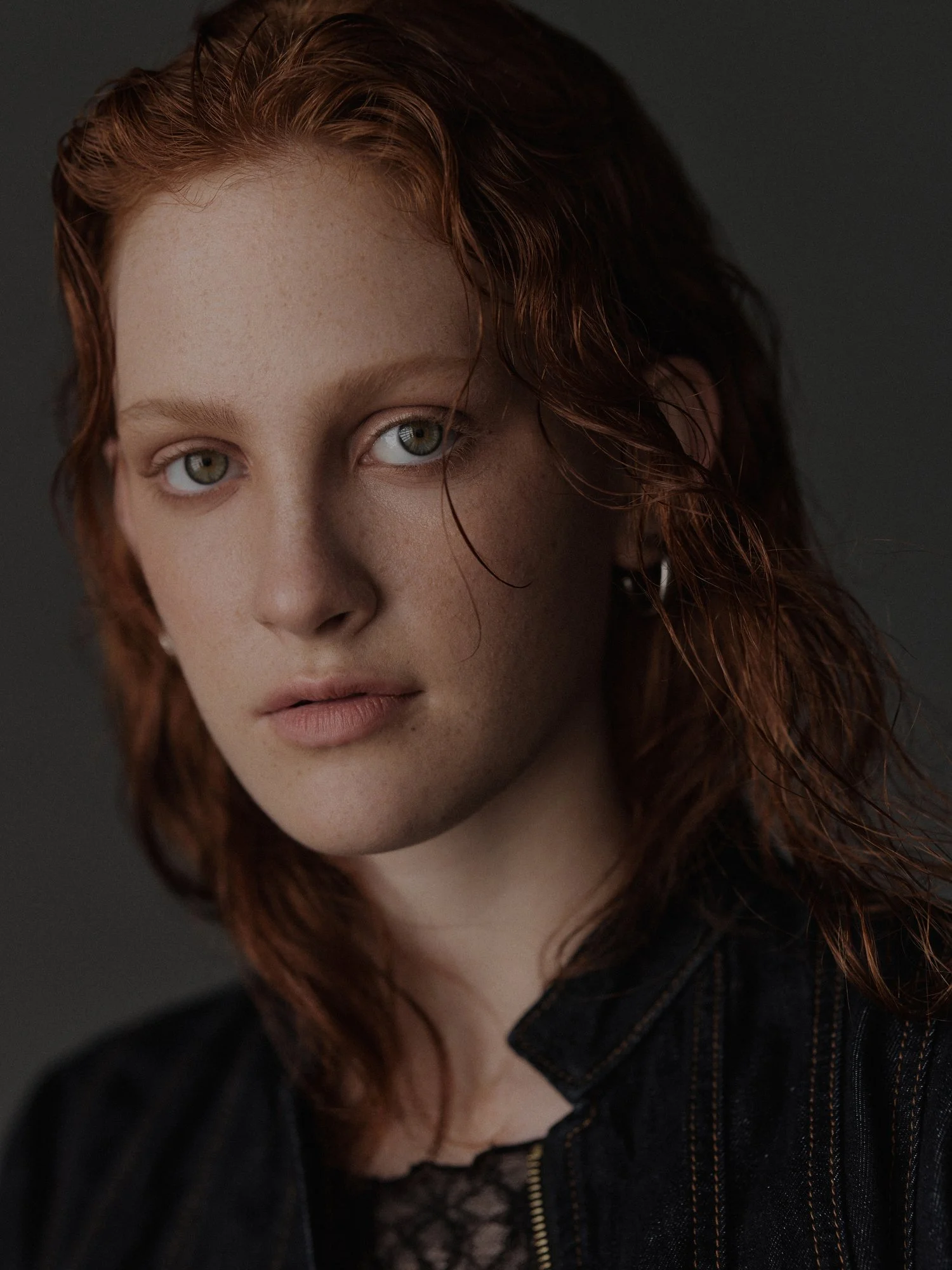 Close-up portrait of a young woman with red curly hair, fair skin, and light hazel eyes, wearing a black jacket with visible stitching.