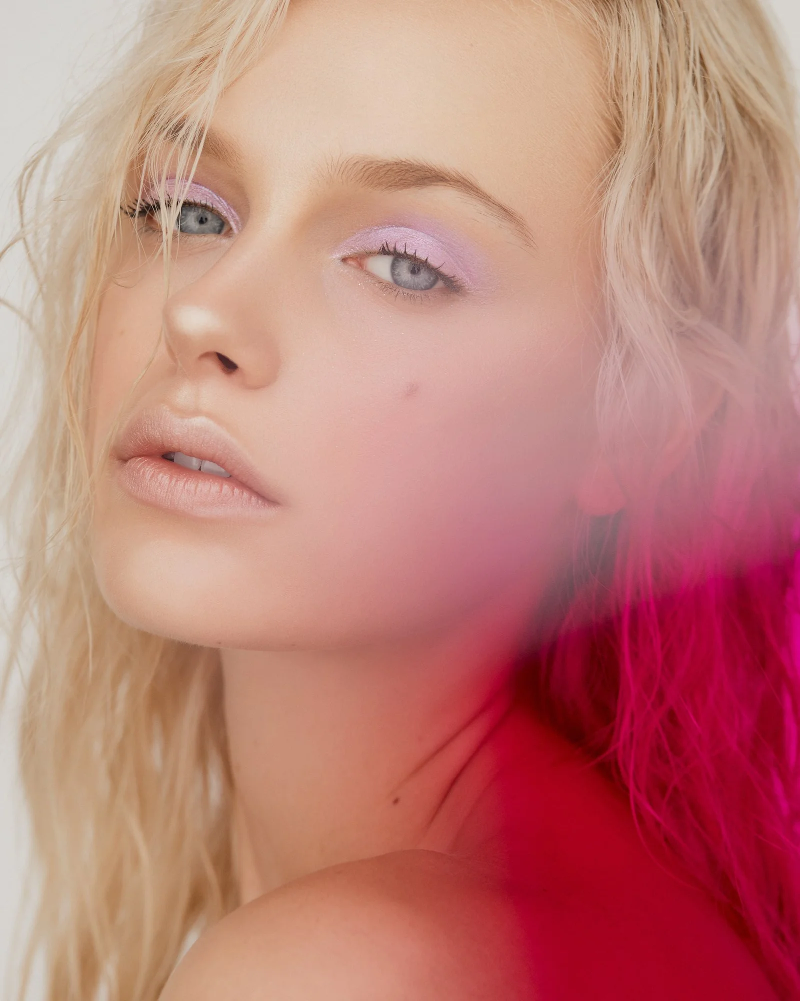 Close-up of a woman's face with platinum blonde hair, purple eyeshadow, and pink feathers on her shoulder.