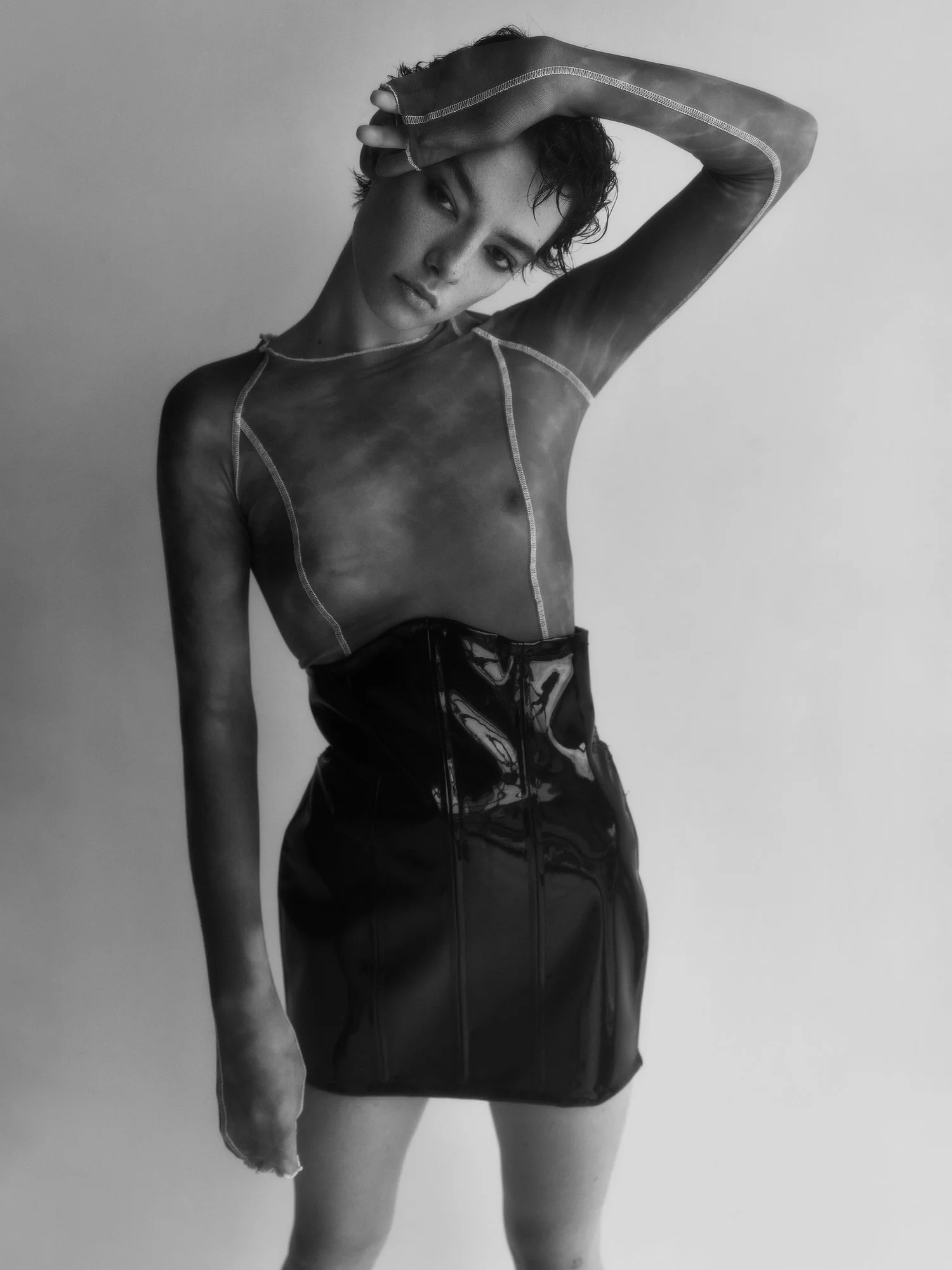 Black and white photo of a woman with short, curly hair wearing a sheer, long-sleeved top and a tight, shiny skirt, posing with one hand on her forehead.
