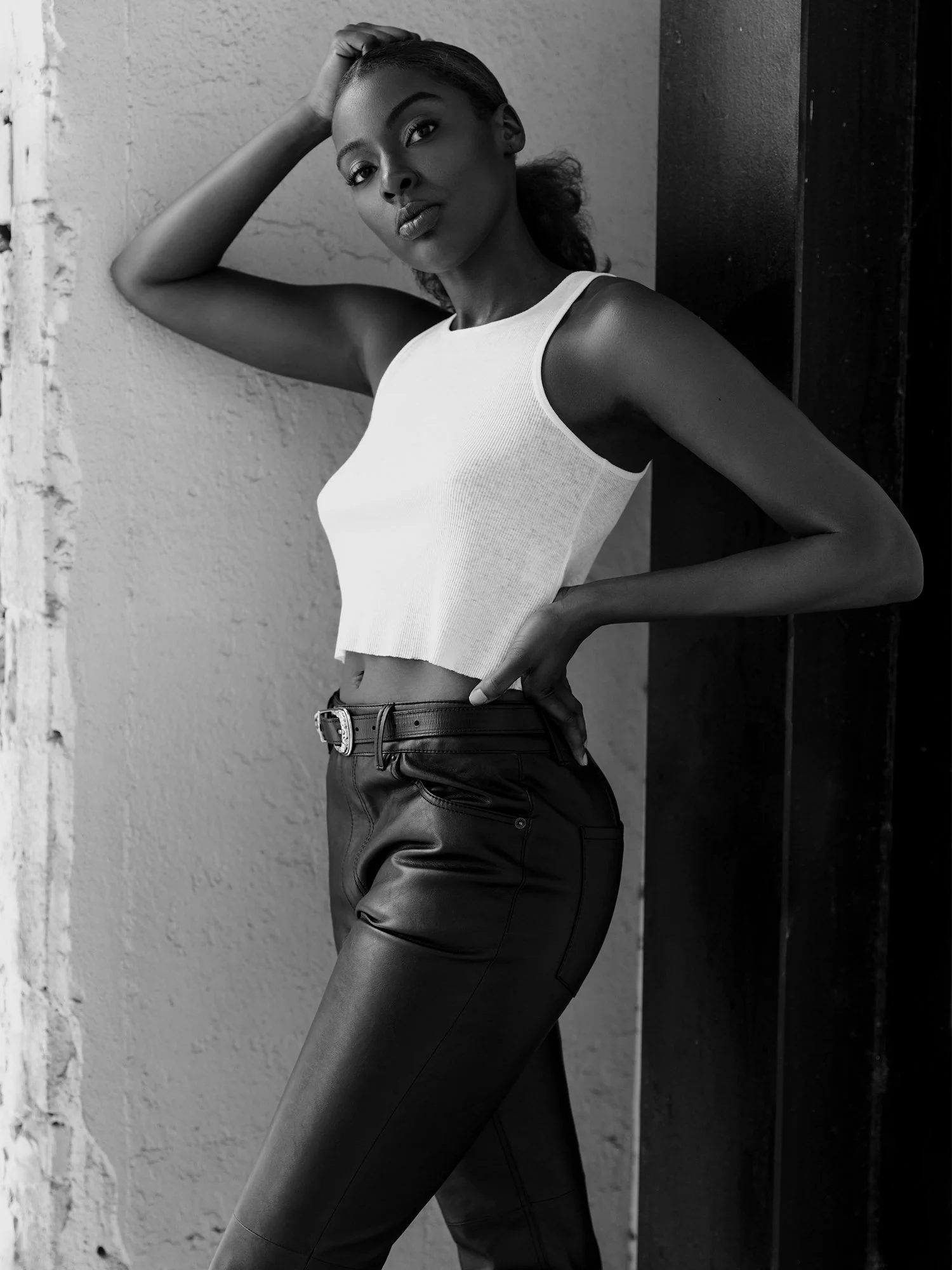 A black-and-white photo of a woman with dark skin, posing with her hand on her head and her other on her hip, standing against a textured wall and a dark vertical surface.