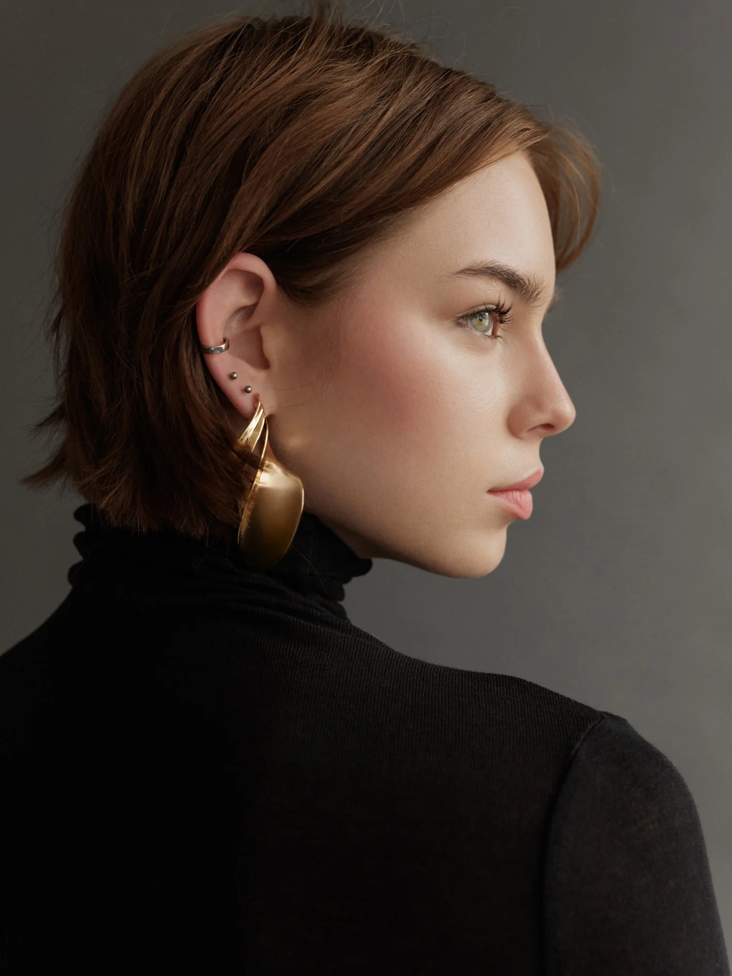 Side profile of a woman with short brown hair, wearing multiple earrings and a black turtleneck, against a gray background.