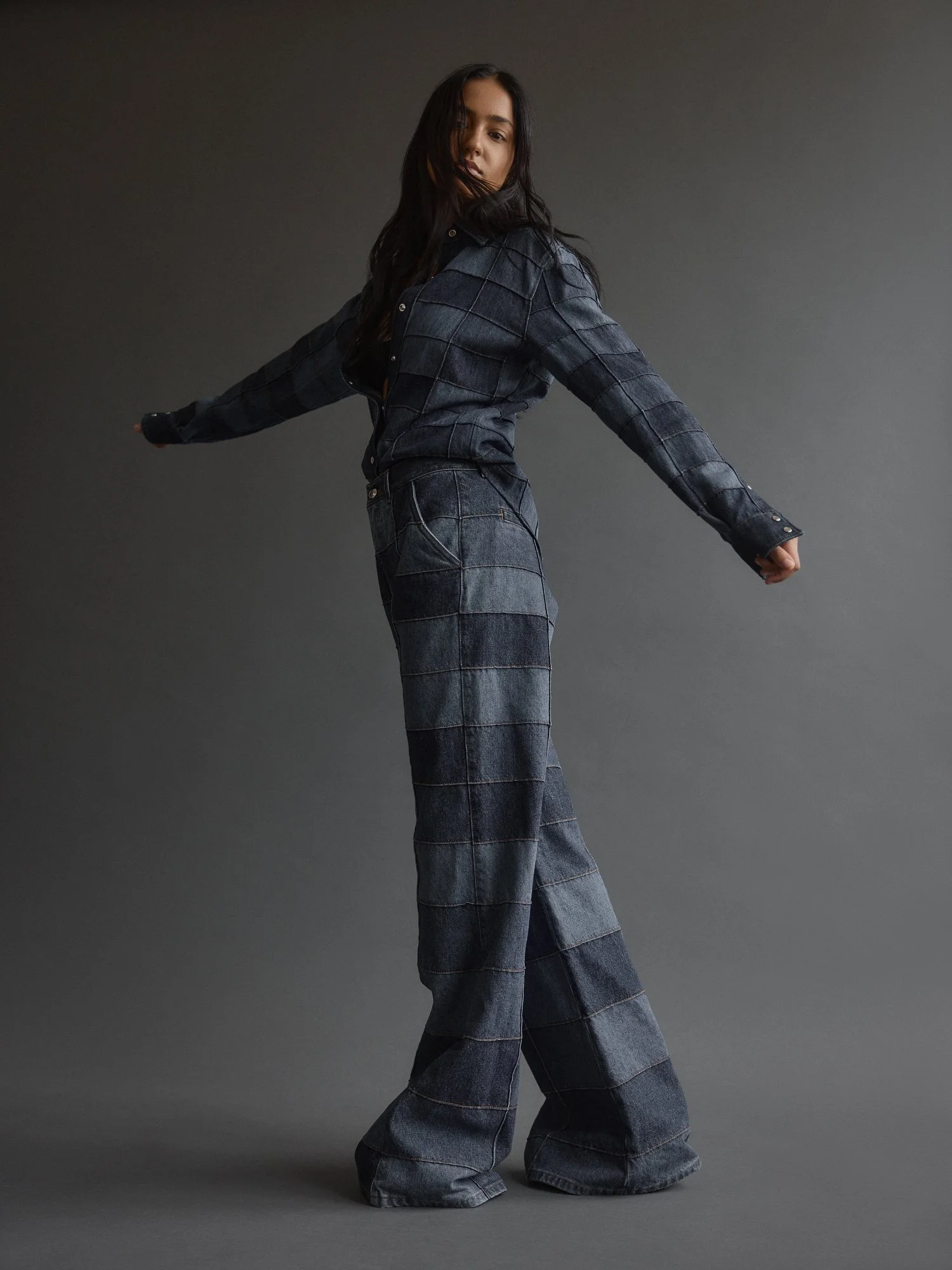 A young woman with long dark hair wearing an oversized dark plaid shirt and wide-leg jeans, standing against a plain gray background.