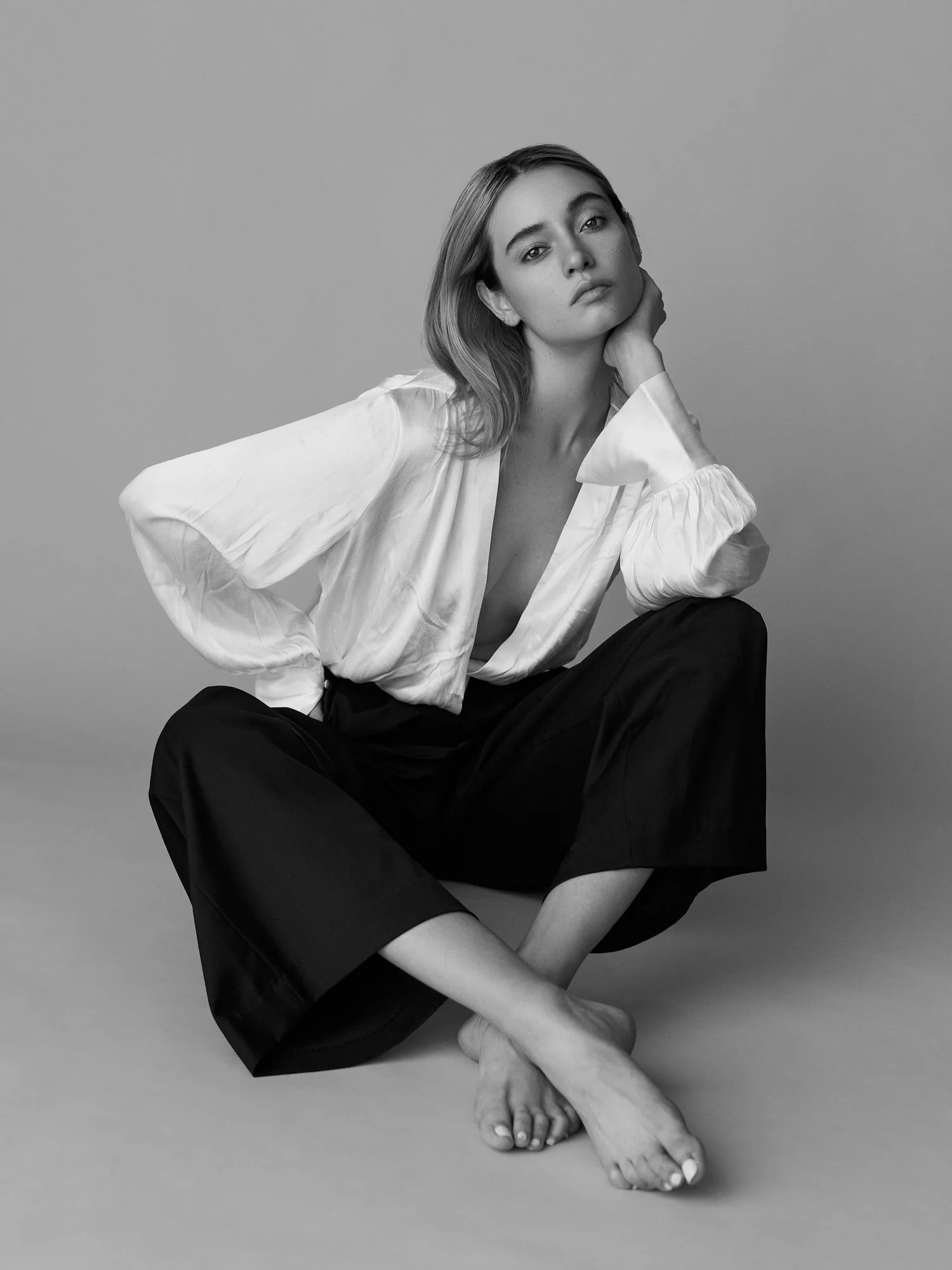 Black and white photo of a woman sitting on the floor, wearing a loose blouse and dark pants, with one hand resting on her face and the other arm resting on her knee, against a plain background.