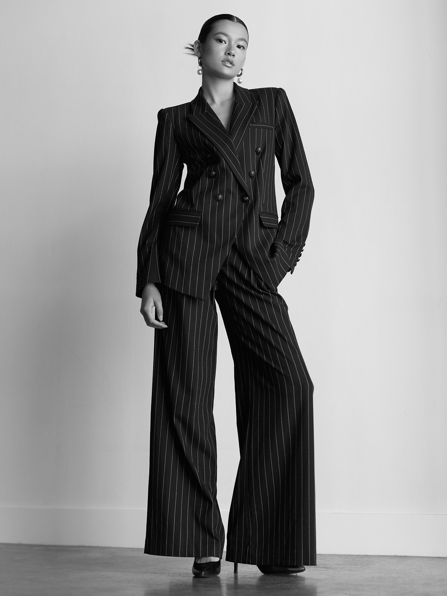 A woman in a pinstripe suit with wide-leg trousers, standing indoors against a plain wall. She has slicked-back hair, wears large pearl earrings, and black high-heeled shoes.