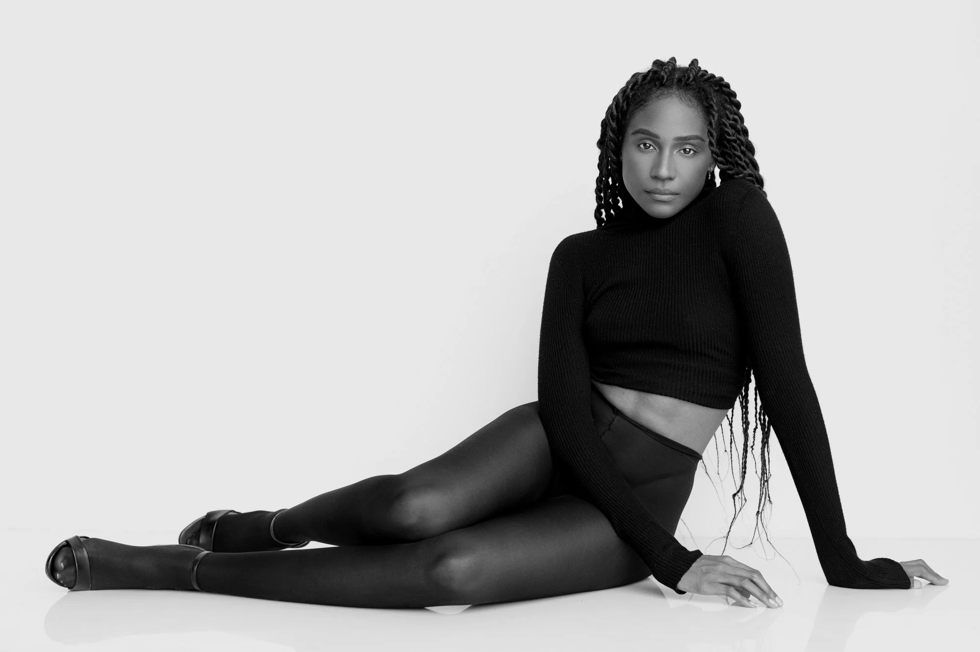 Black woman with long dreadlocks, dressed in black, sitting on floor with legs extended, in front of a plain white background in a studio setting.