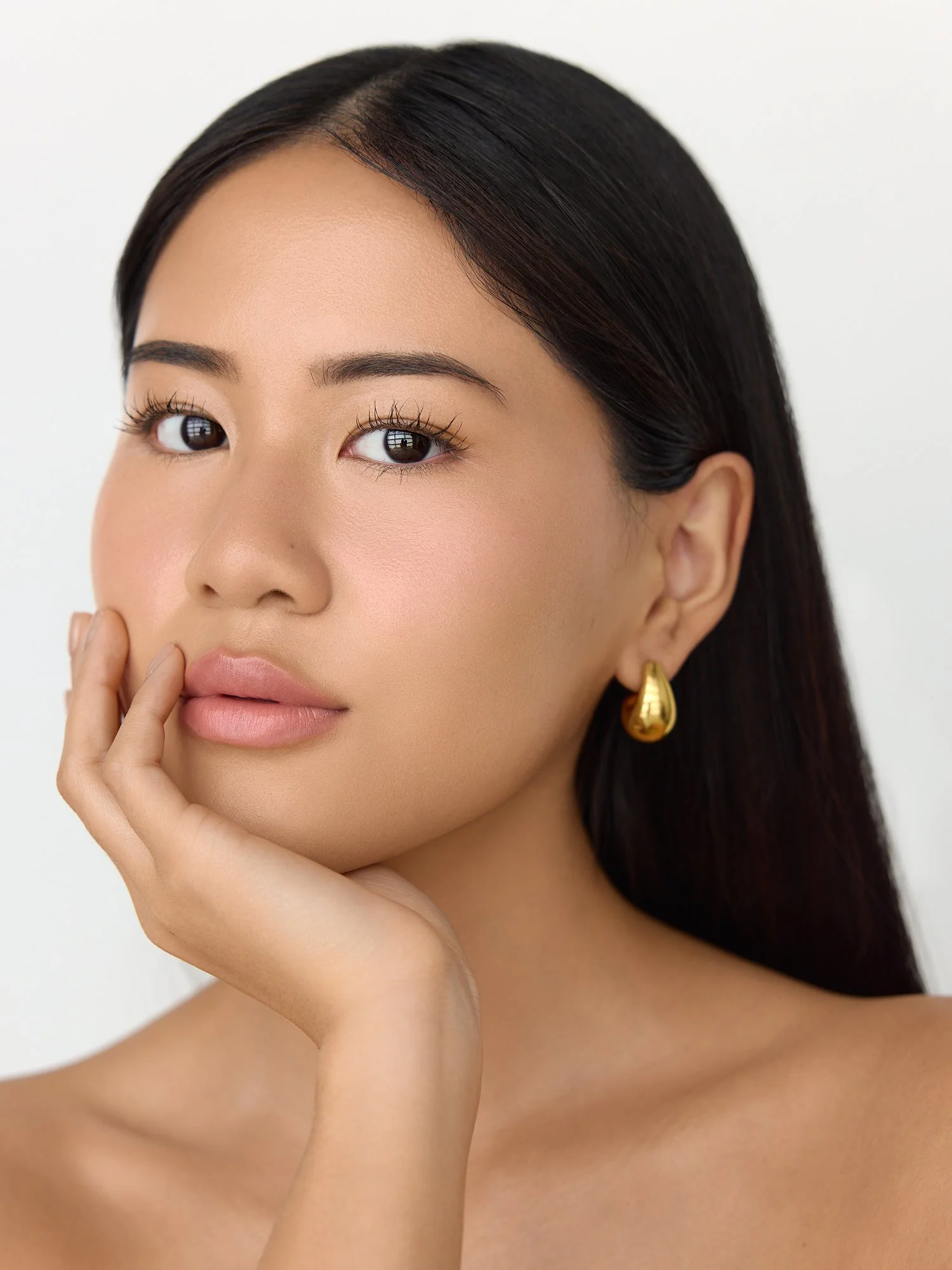 Close-up of a woman with smooth skin, dark hair, and gold teardrop earring, touching her face with her hand.