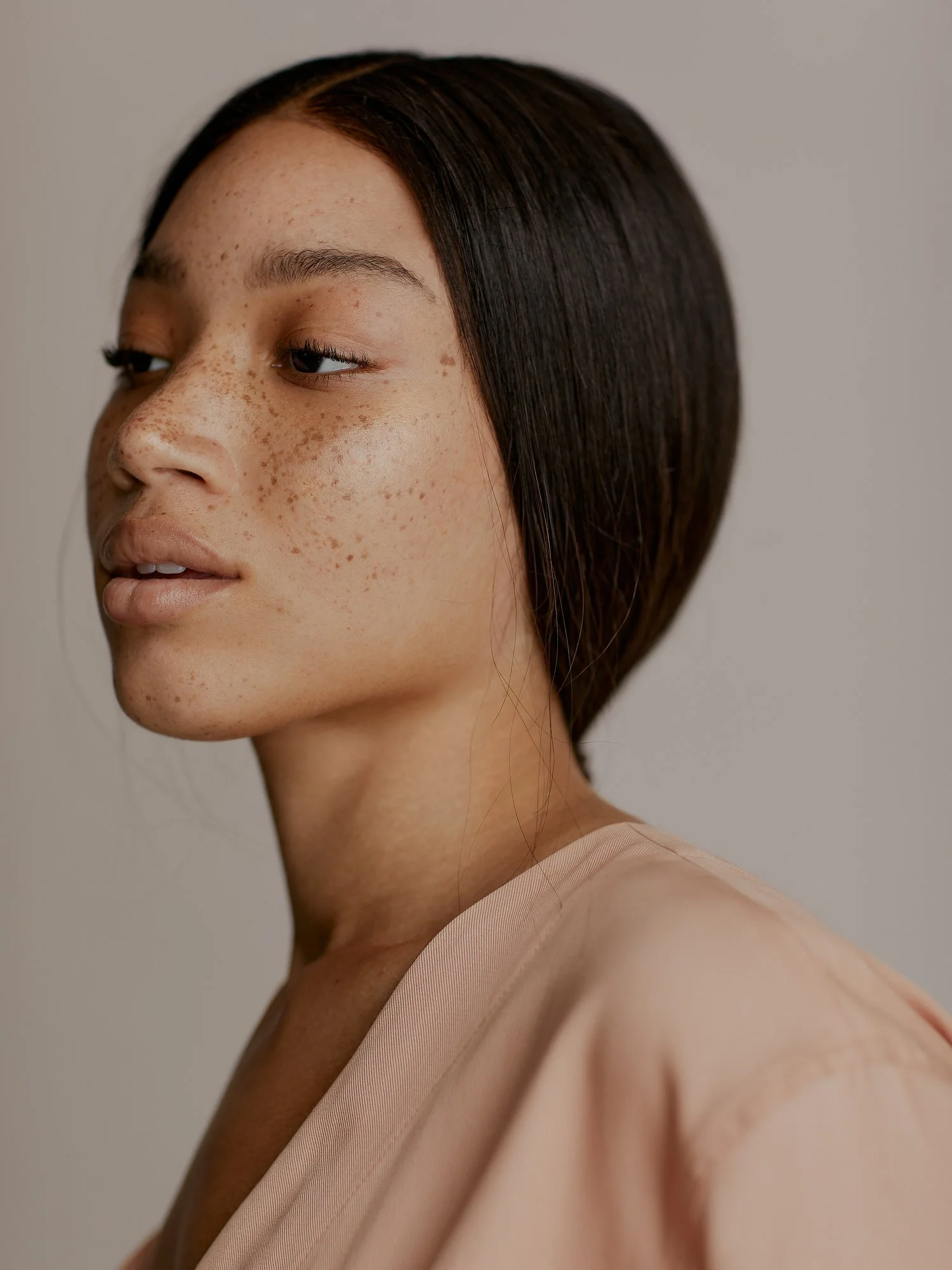 A close-up of a woman with dark brown hair, light skin, and freckles, wearing a soft pink top, looking slightly to her left against a plain light background.
