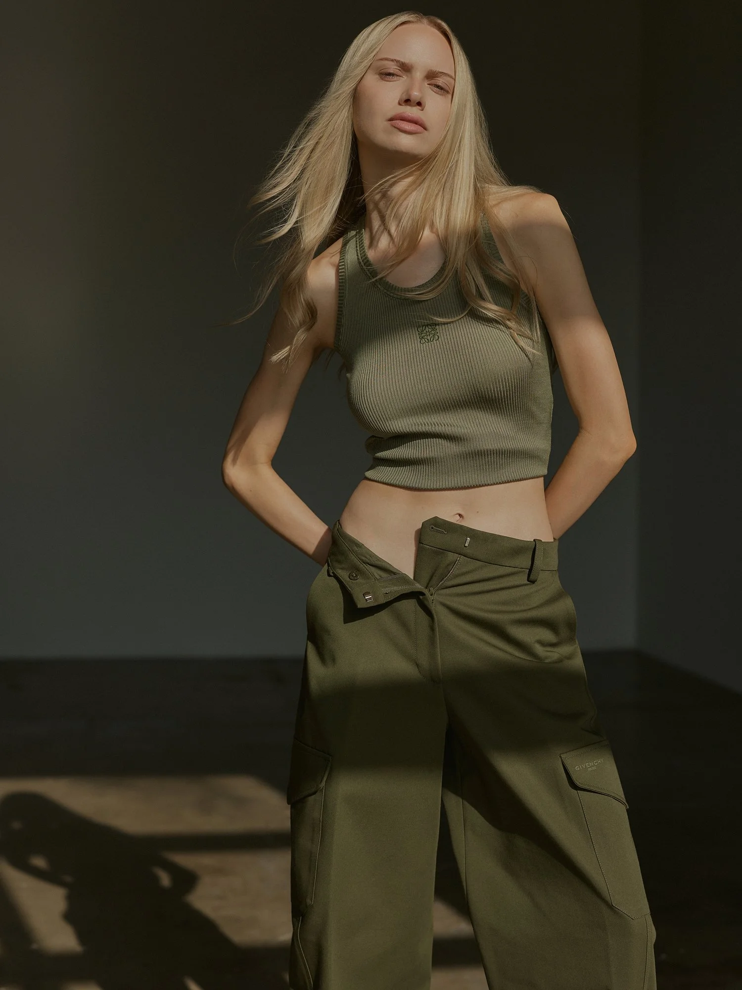 A woman with long blonde hair in a green tank top and olive cargo pants stands with one hand on her hip against a dark background.