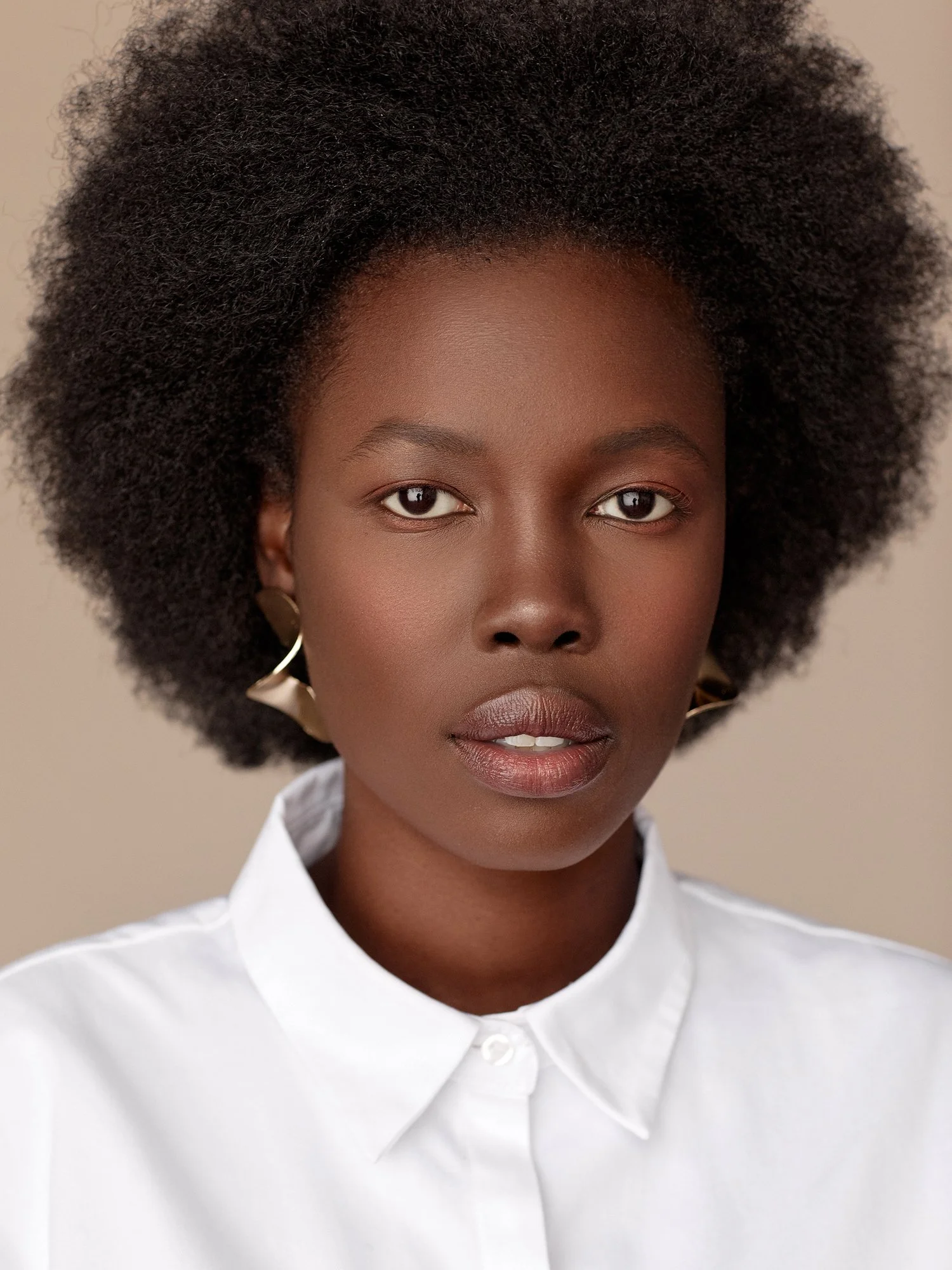 Close-up portrait of a woman with natural afro hair, wearing a white shirt and gold earrings, against a neutral background.