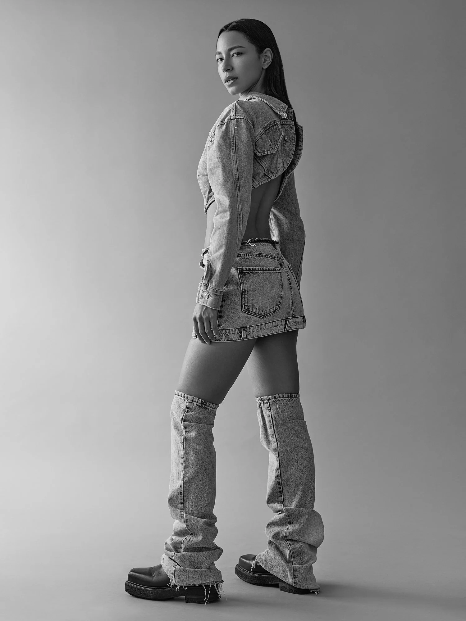 Black and white photo of a woman wearing a denim jacket and jeans, standing with her body turned to the side, looking at the camera with a serious expression.