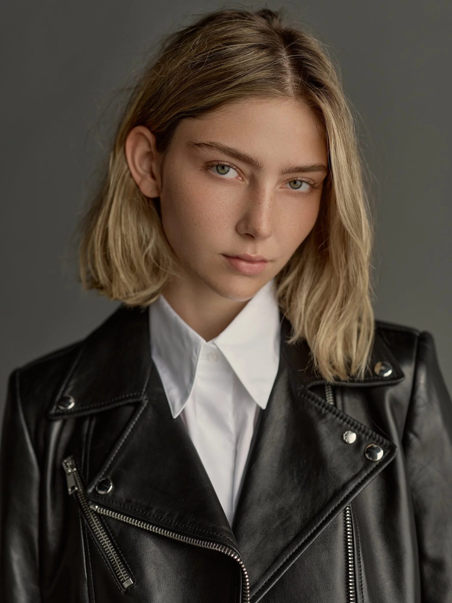 Close-up of a young woman with blonde hair and green eyes wearing a white collared shirt and a black leather jacket with silver zippers.