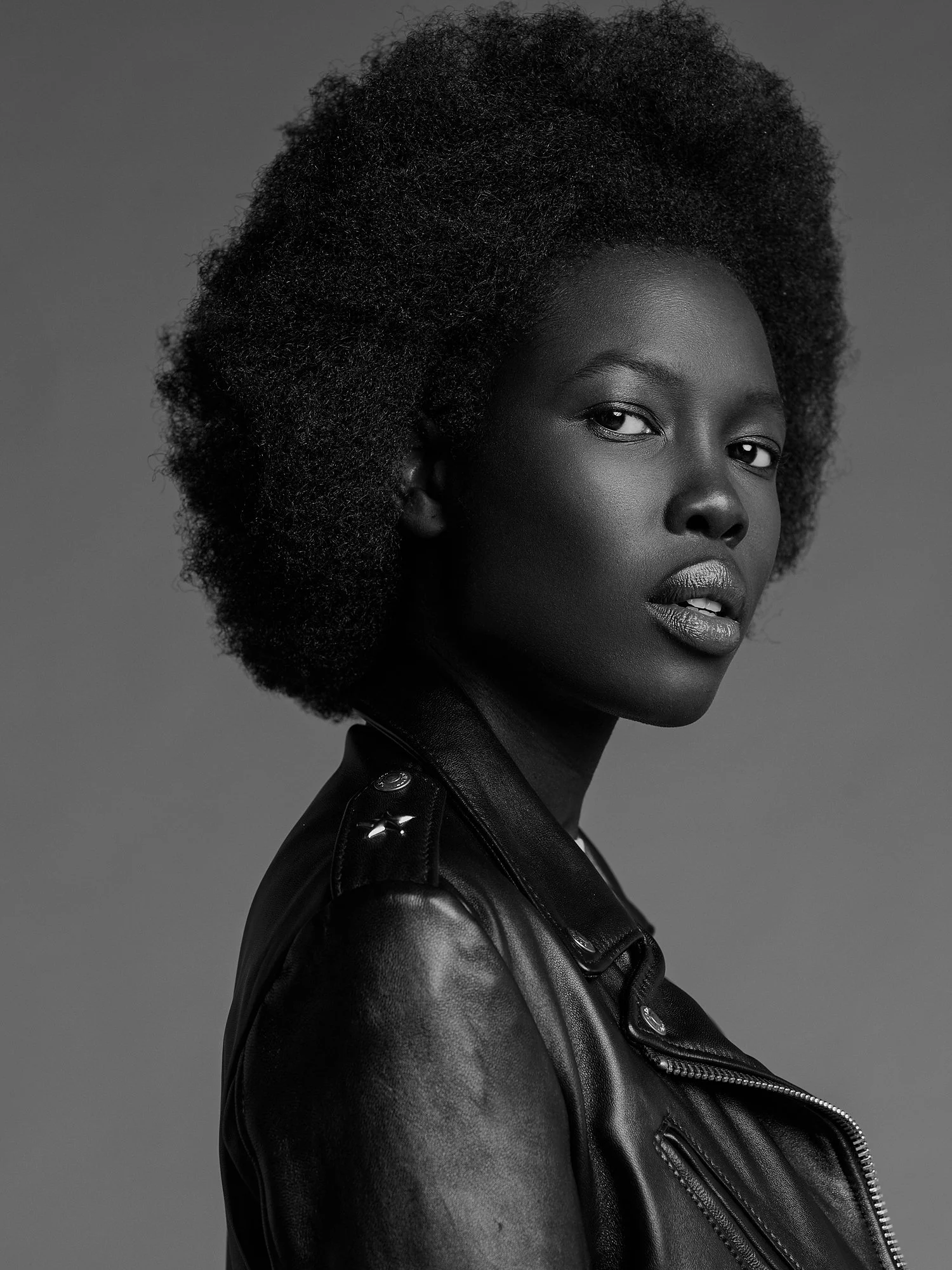 A Black woman with natural hair wearing a black leather jacket, looking at the camera against a gray background.