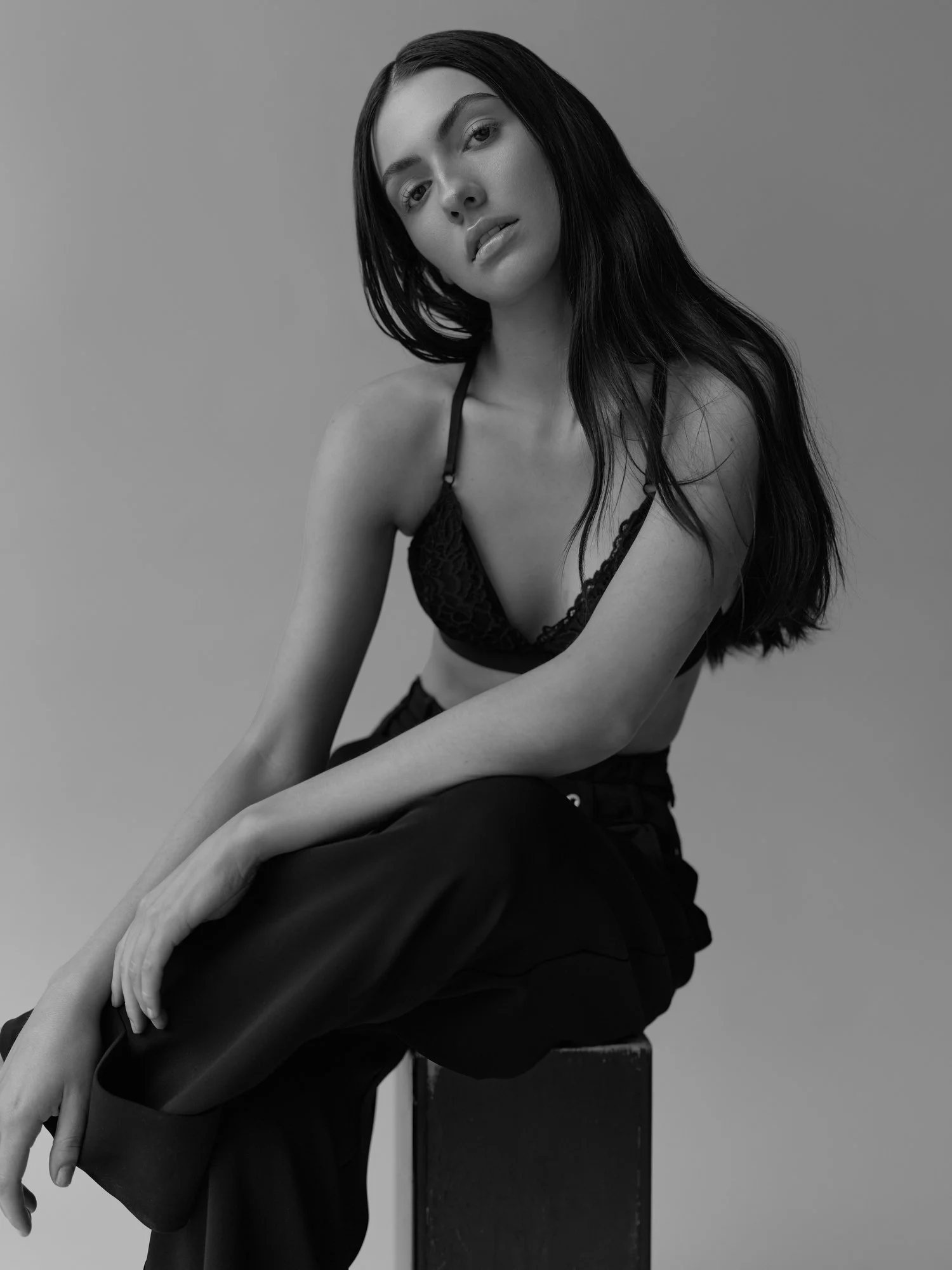 Black and white photograph of a woman with long black hair, wearing a lace top, sitting on a wooden block, with her hands resting on her knees, against a plain background.