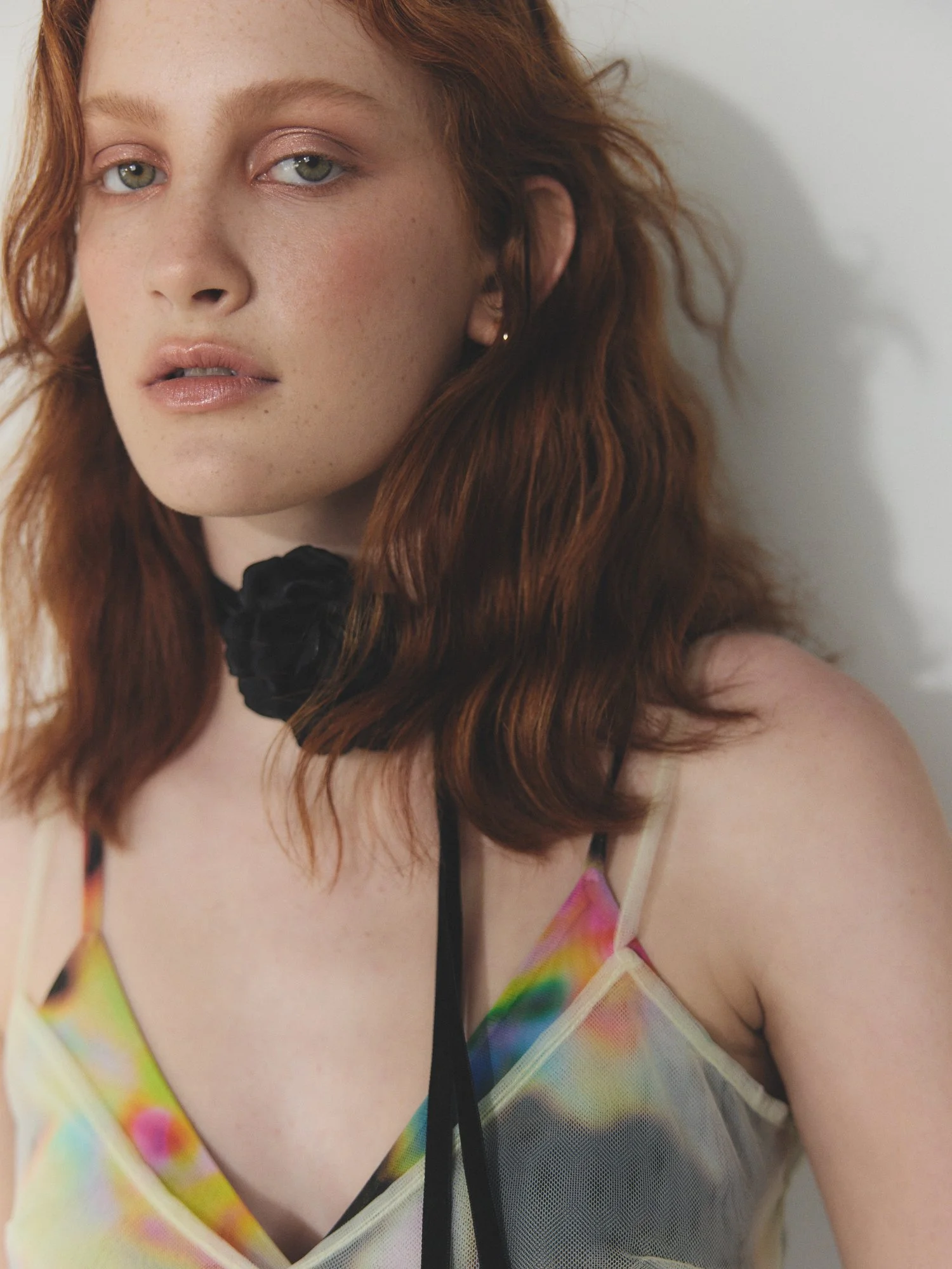 Close-up of a young woman with red hair, wearing eyeshadow and a colorful rainbow-patterned top, facing the camera with a neutral expression.