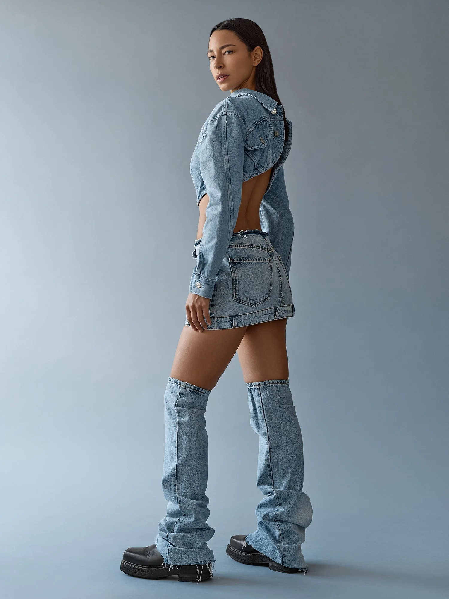 Young woman with long dark hair posing against a plain gray background, wearing a unique denim jacket with open back design, high-waisted jeans, and black platform boots.