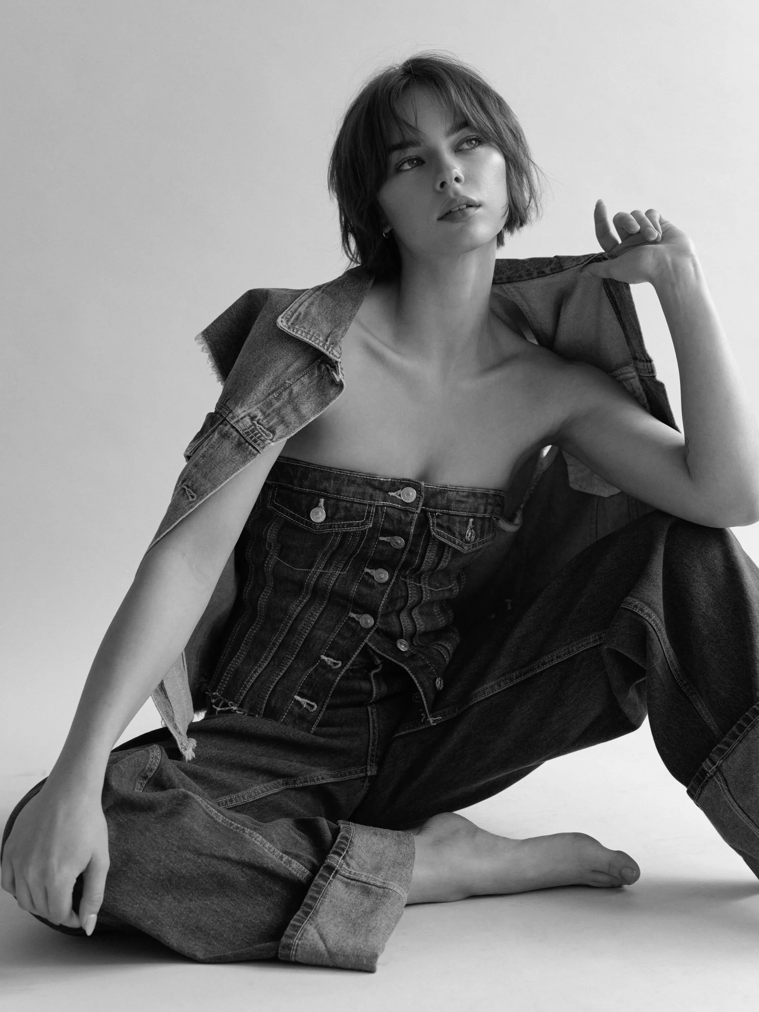 Black and white photo of a woman with short wavy hair, wearing a denim jacket and jeans, sitting on the floor with one leg crossed under the other, looking upward and to the right.