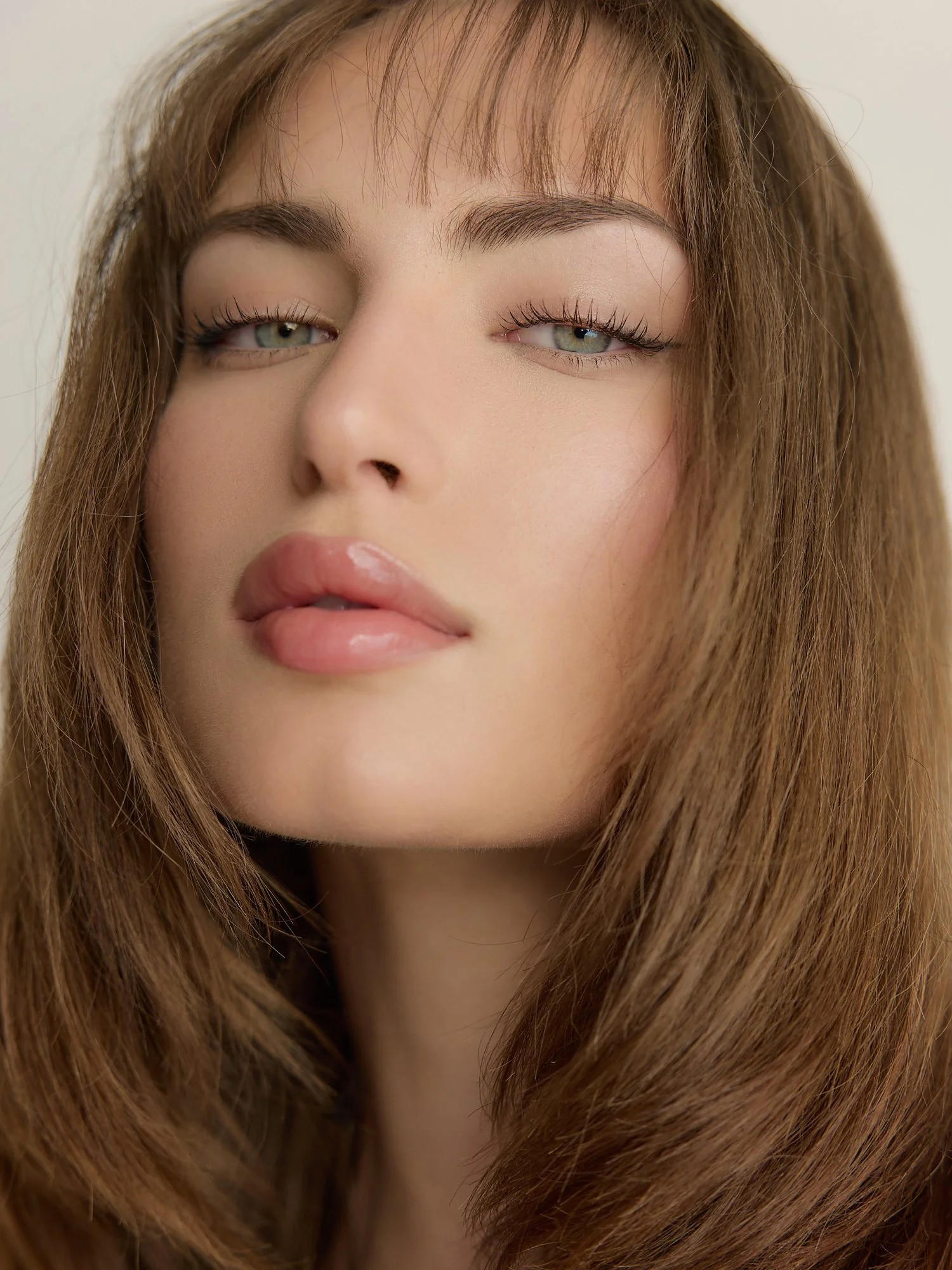 Close-up of a woman with light green eyes, light makeup, and long brown hair with bangs, looking softly at the camera.