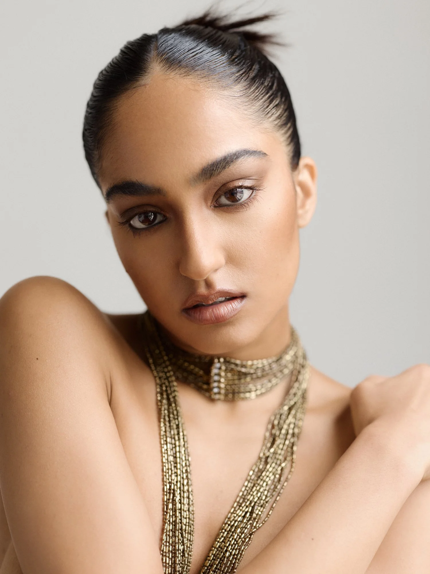 Close-up of a woman with sleek dark hair in a tight, polished bun, wearing multiple gold chain necklaces, with a neutral background.