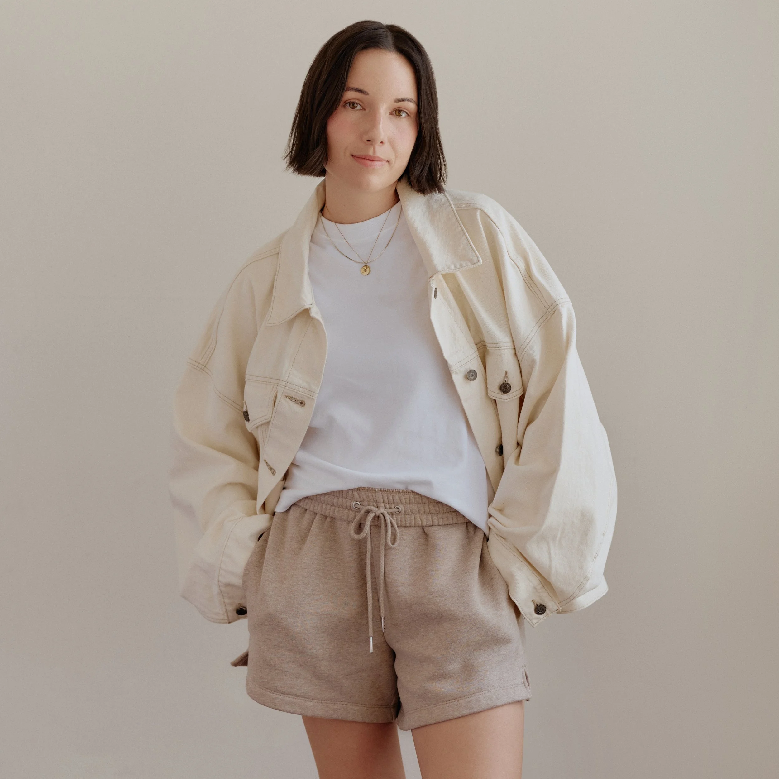 A woman with short dark hair, wearing a white t-shirt, beige shorts, and a beige jacket, stands against a plain, light-colored background.