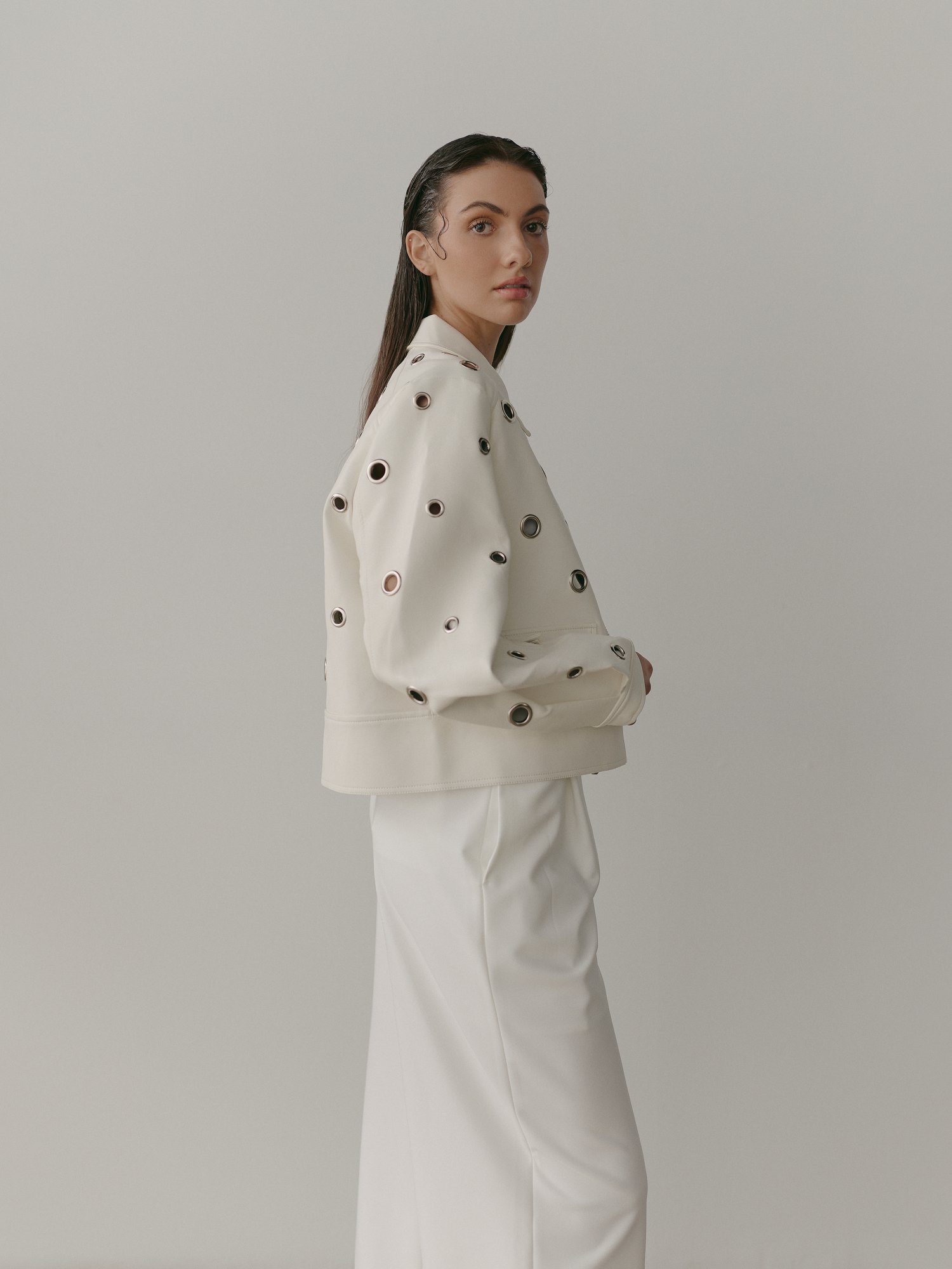 A woman with wet hair wears a white jacket with metallic eyelet details and white pants, standing against a plain gray background.