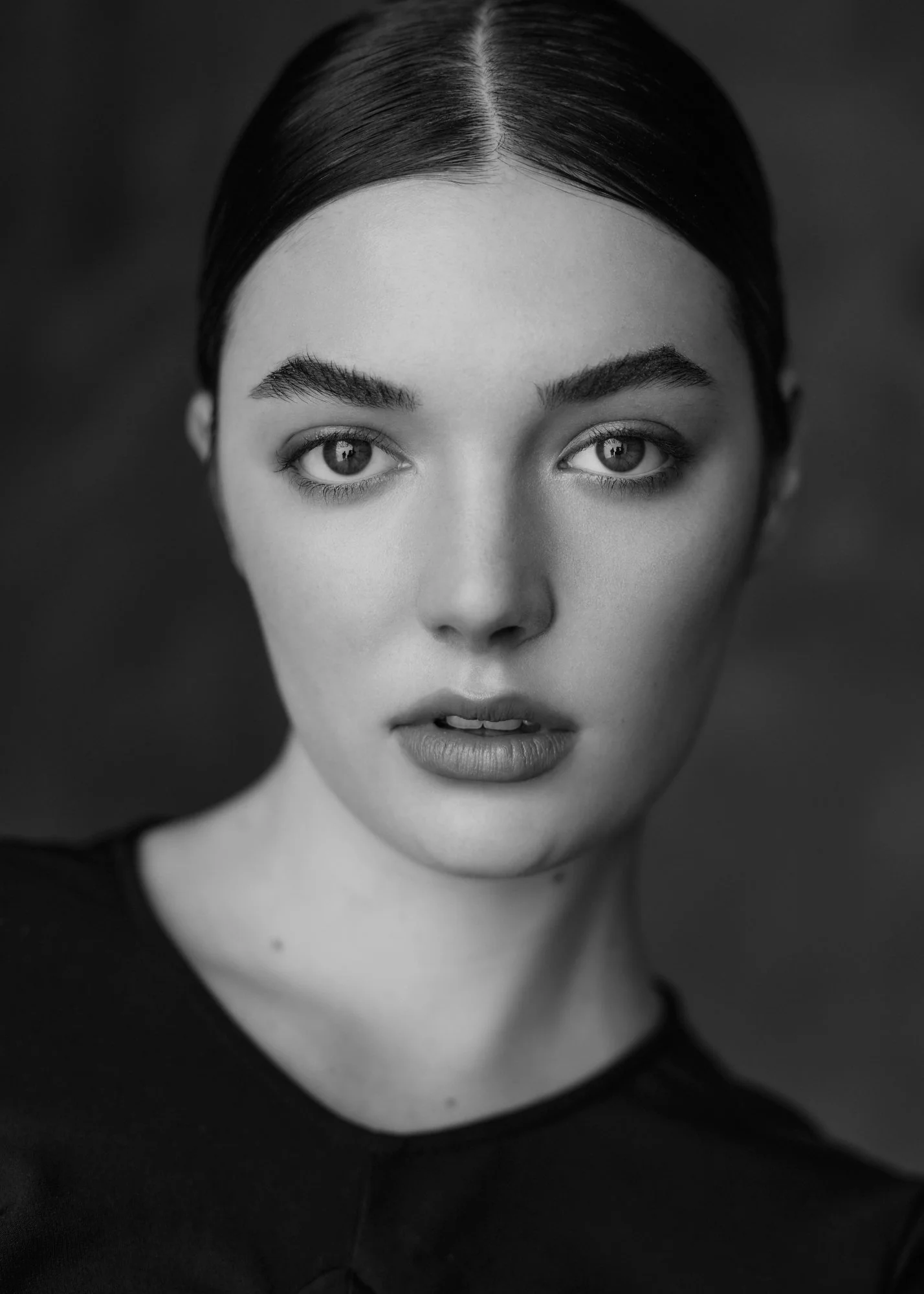 Close-up black and white portrait of a young woman with dark hair styled slicked back, intense gaze, full lips, and defined eyebrows, wearing a dark top.