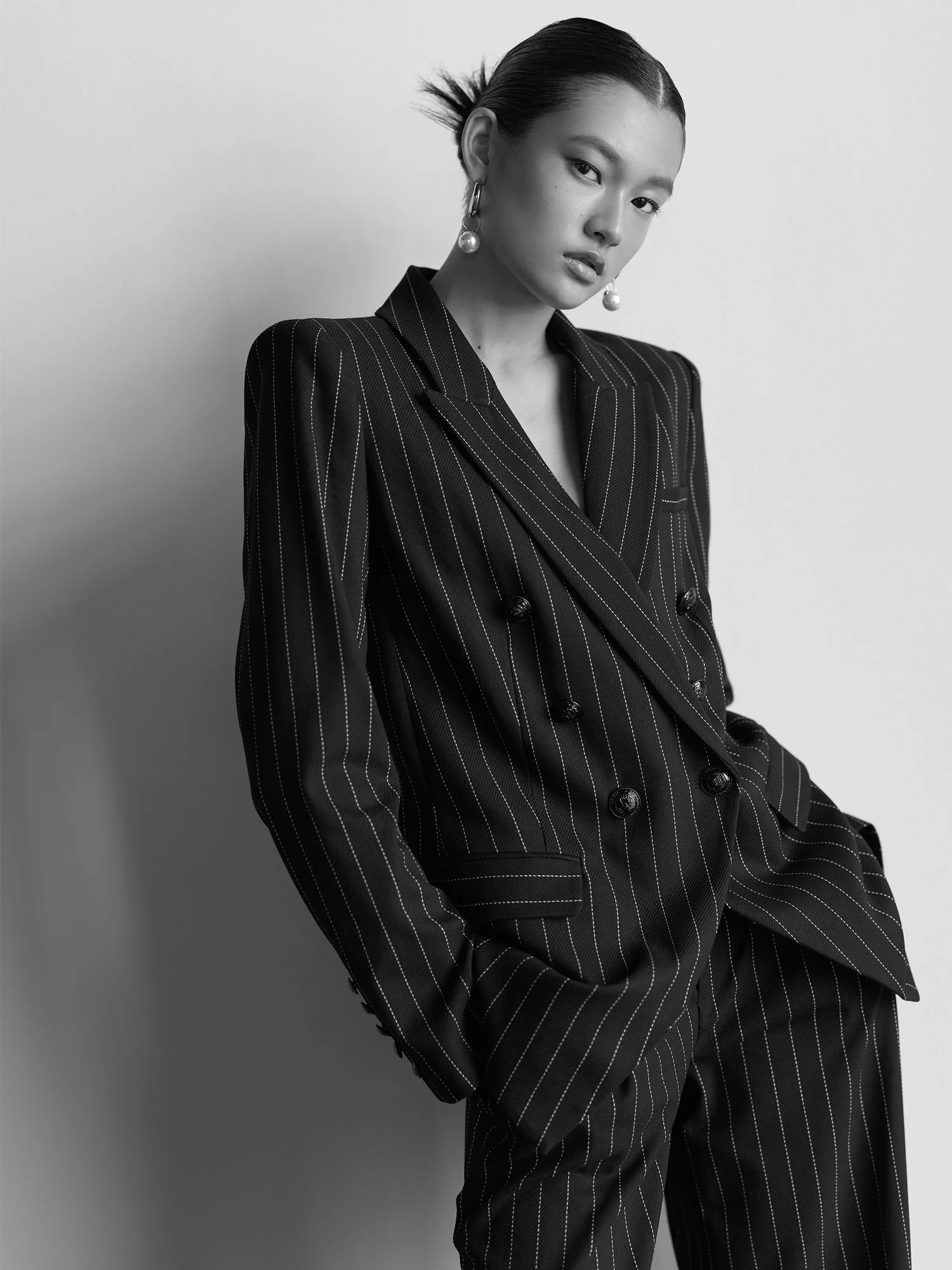 A black and white portrait of a woman with slicked-back hair wearing a pinstripe blazer and large pearl earrings, looking at the camera with a confident expression.