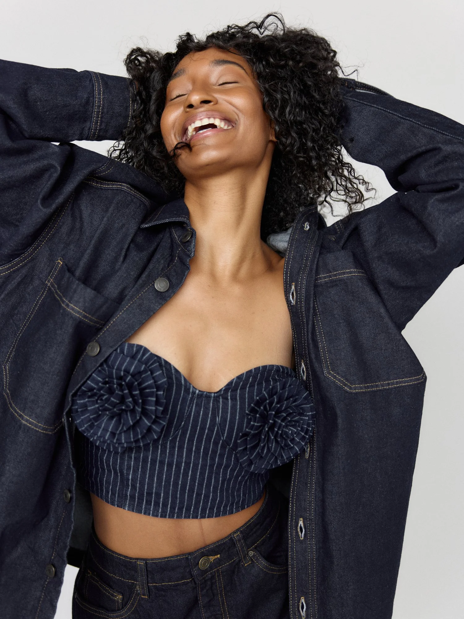 A woman with curly black hair smiling with eyes closed, wearing a denim jacket and matching denim shorts, against a plain white background.