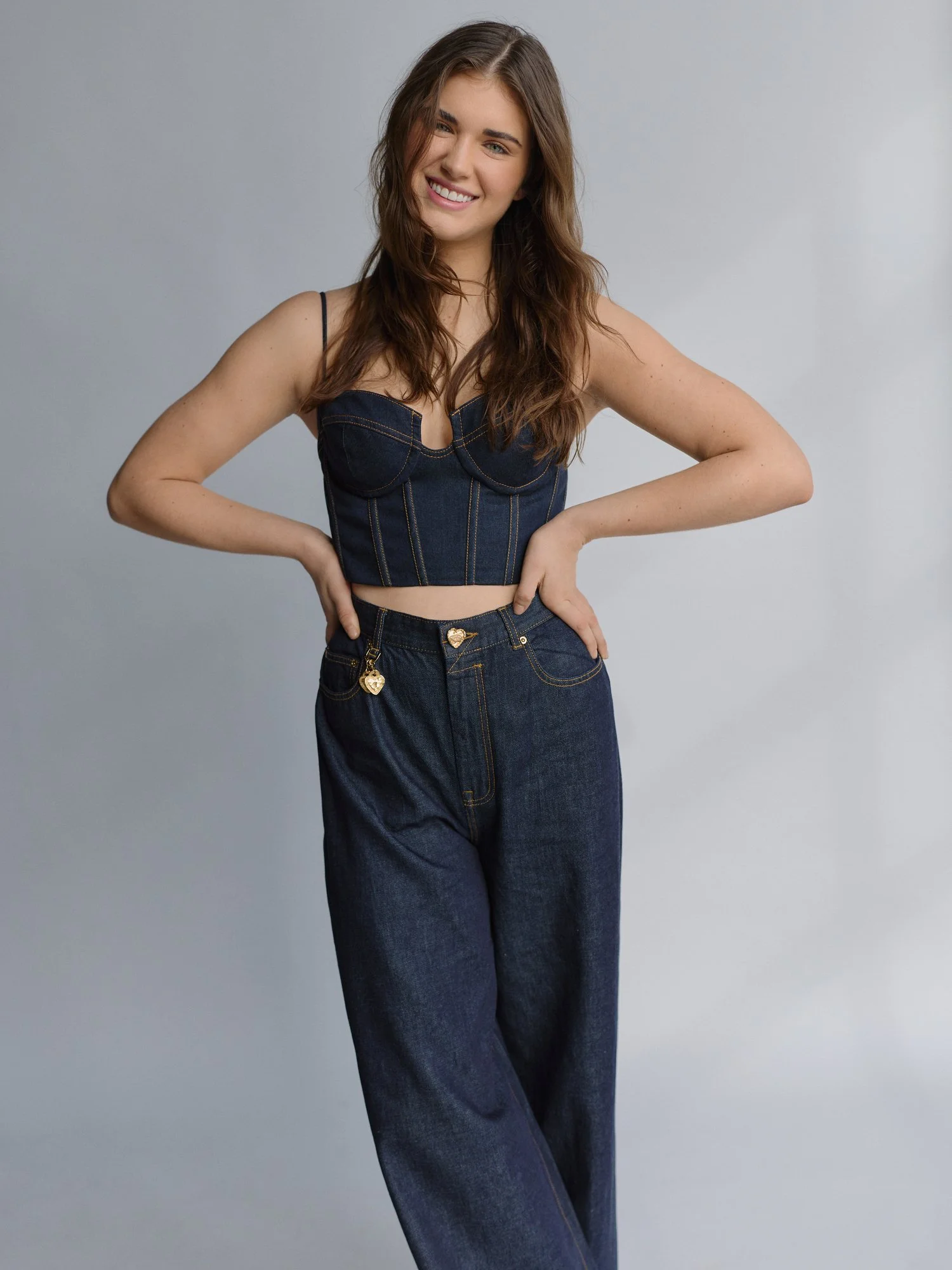 Young woman with long brown hair smiling, wearing a dark denim crop top and matching high-waisted jeans, standing with hands on hips against a light gray background.