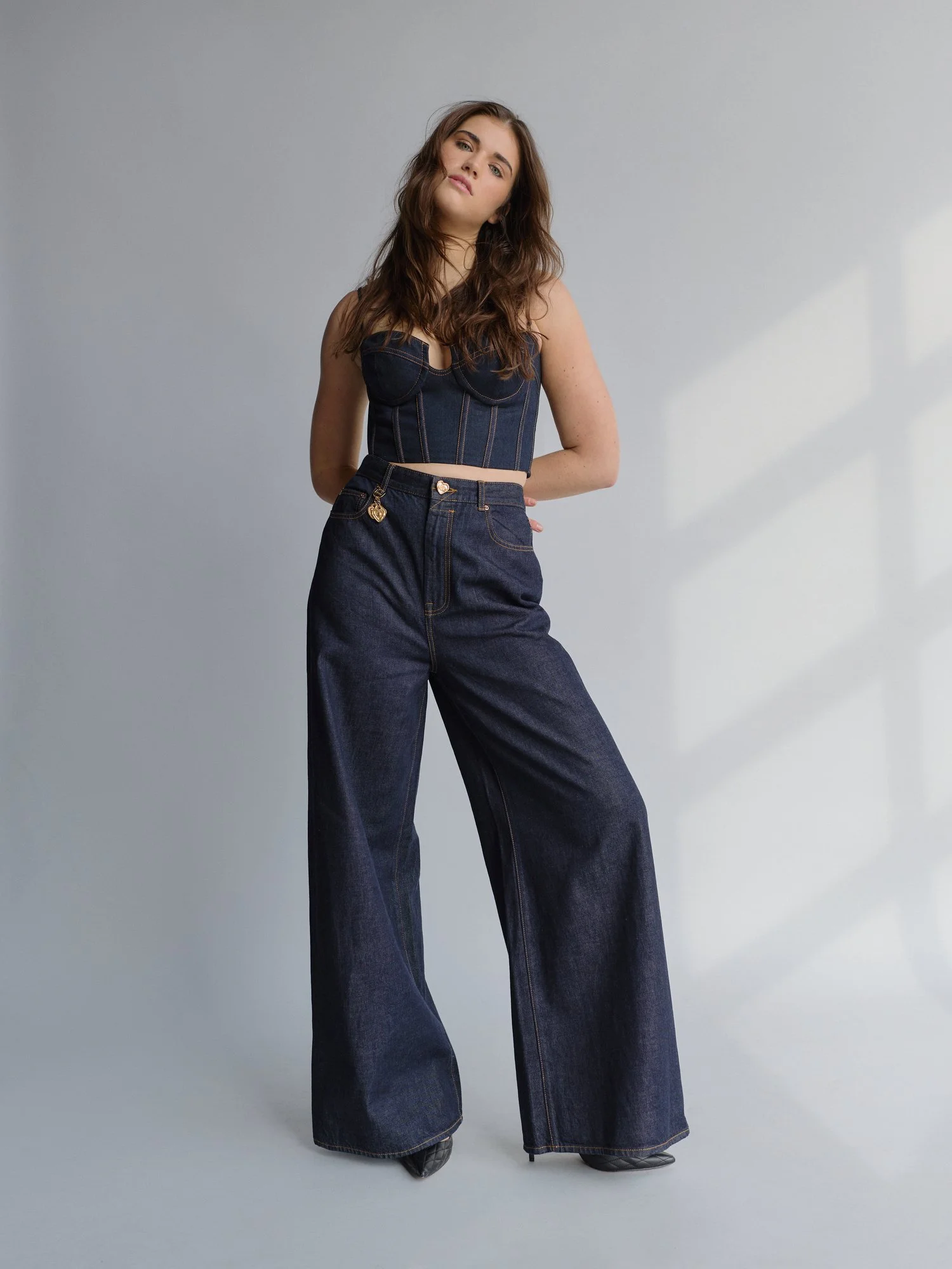 A young woman with long wavy brown hair wearing a dark denim corset top and high-waisted wide-leg jeans standing against a plain light gray background.