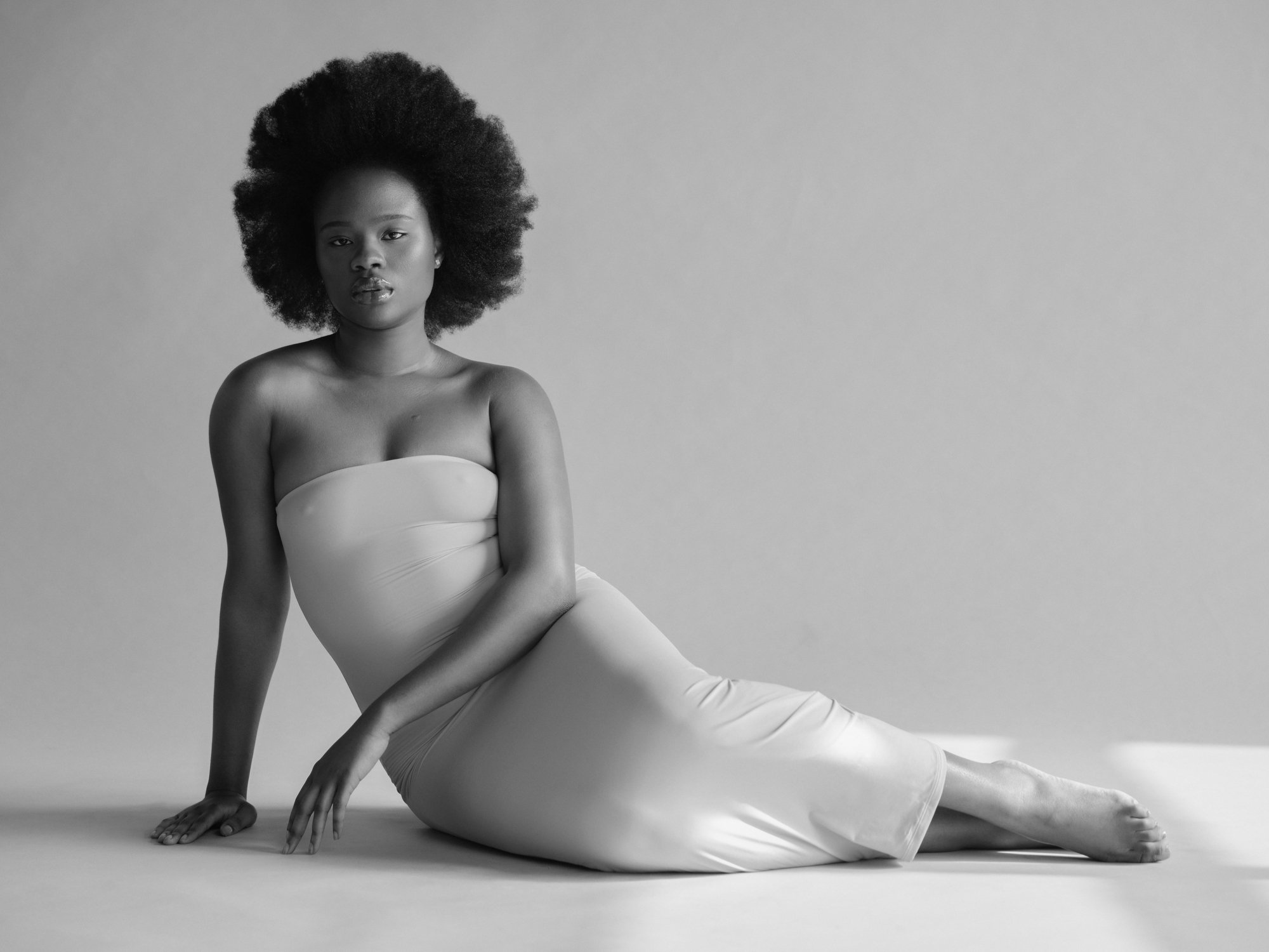 Black and white photo of a woman with natural hair, sitting on the floor with one arm supporting her and the other hand on her knee, wearing a strapless top and pants.